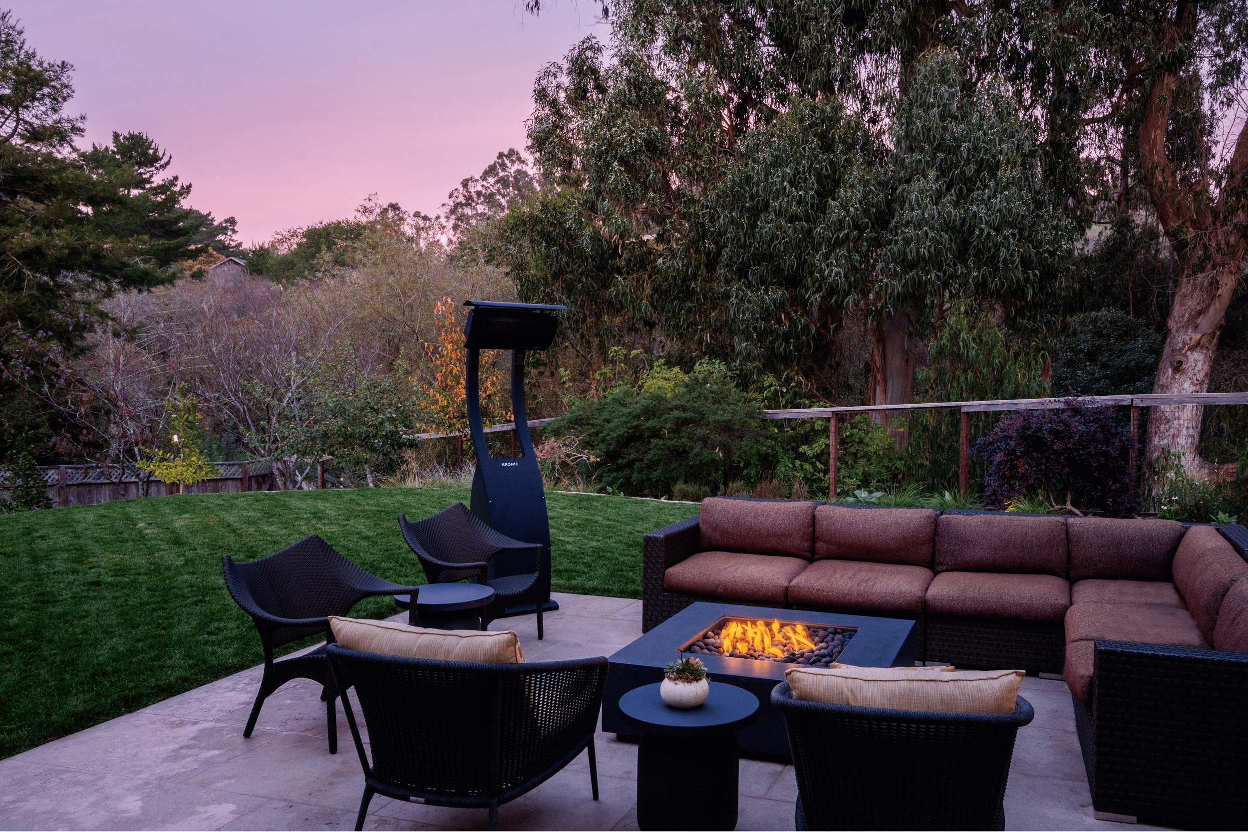 Outdoor patio with a fire pit, chairs, a sofa, and lush trees at sunset.