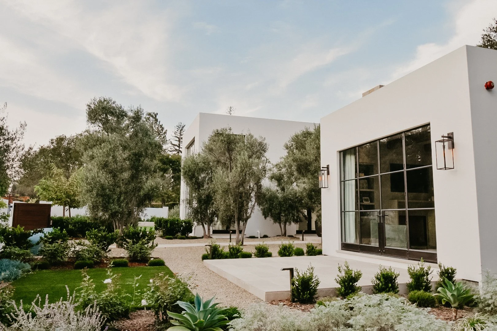 Modern white house with large glass windows, surrounded by landscaped garden with greenery and trees, under a partly cloudy sky.