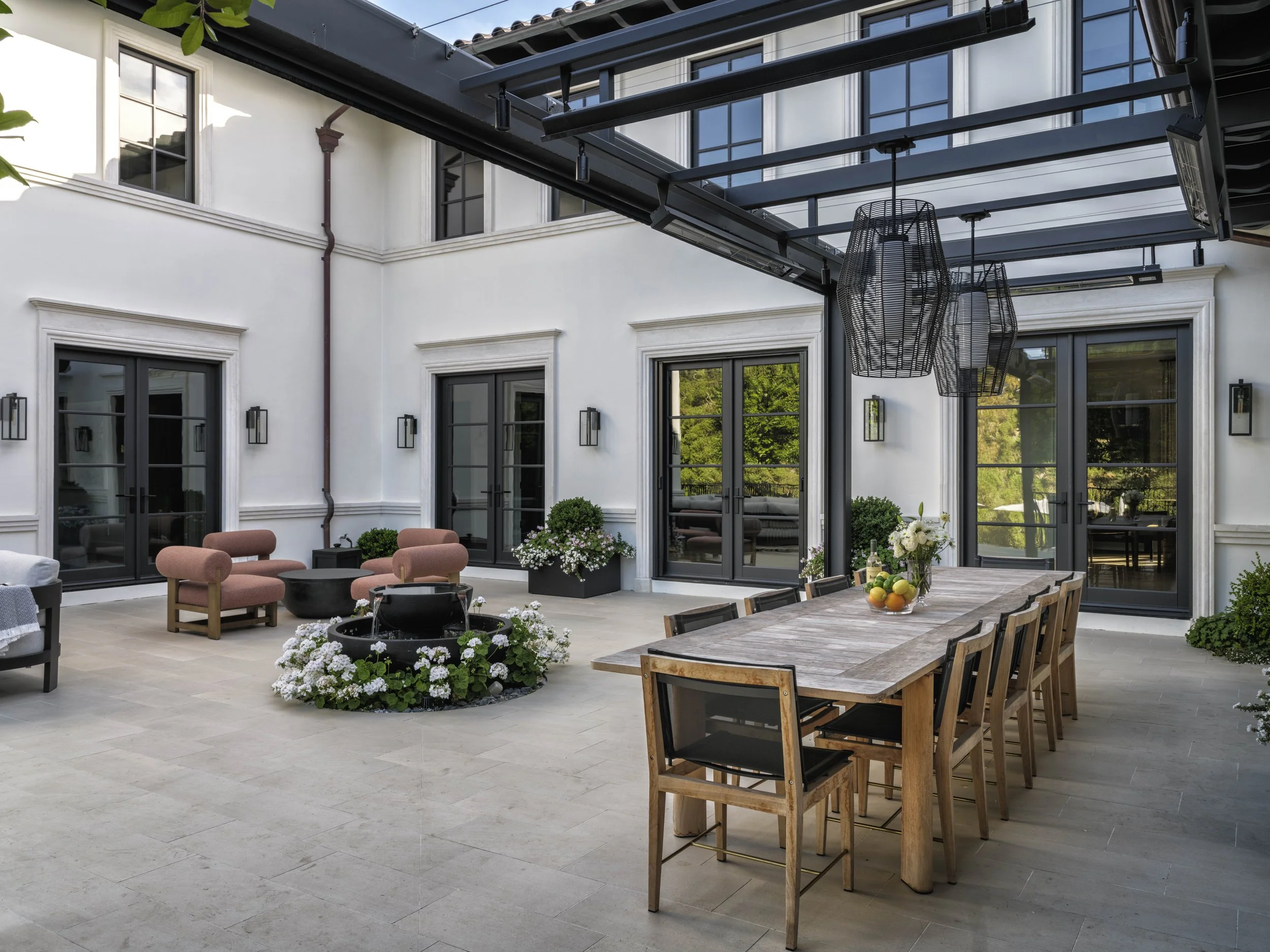 An outdoor patio with a wooden dining table, black chairs, pink lounge chairs, a fire pit, potted plants, and hanging lamps, enclosed by white walls and black-framed glass doors and windows.