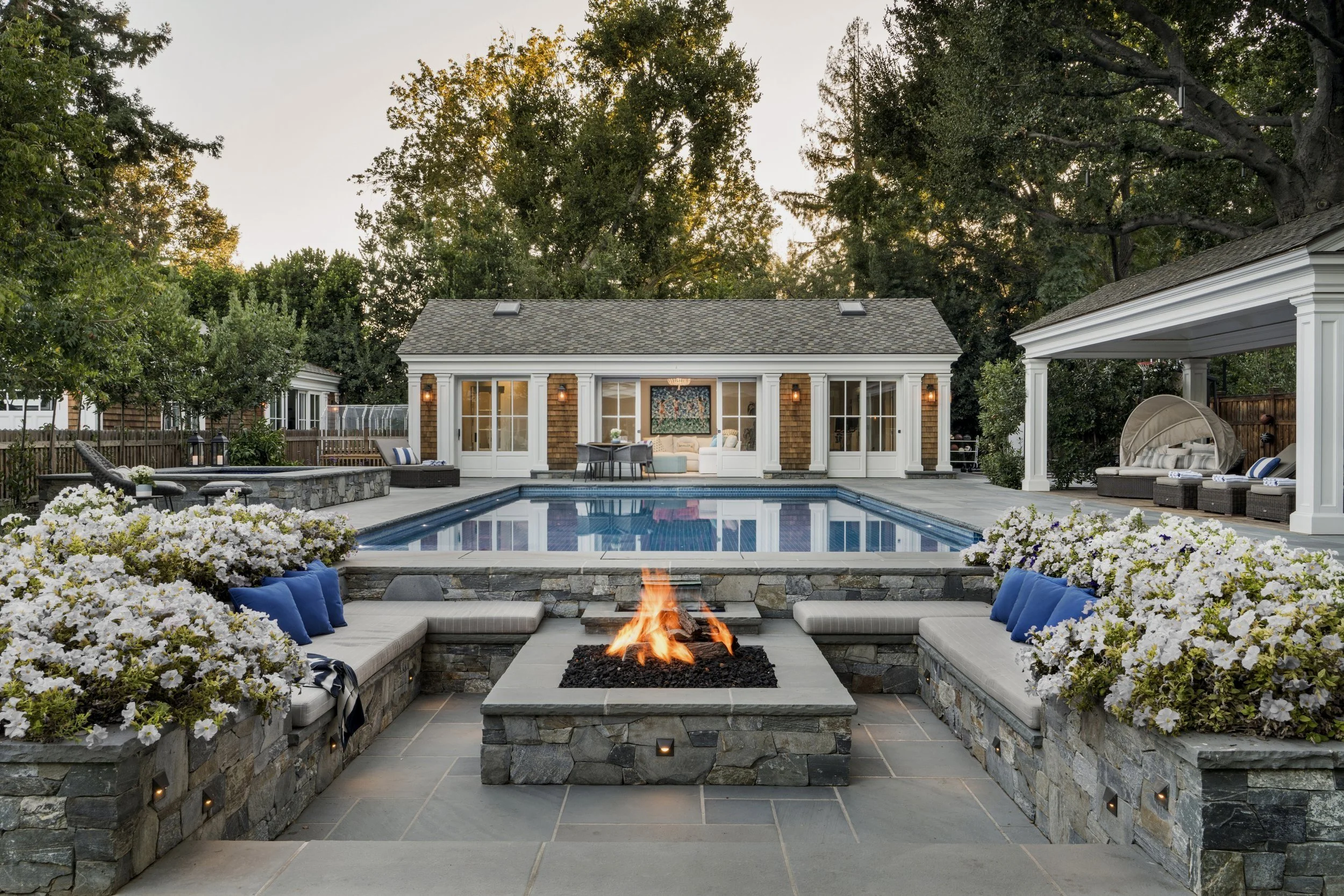 A backyard with a swimming pool, stone fire pit with flames, and outdoor seating areas, surrounded by flowering plants and trees, with a house in the background.