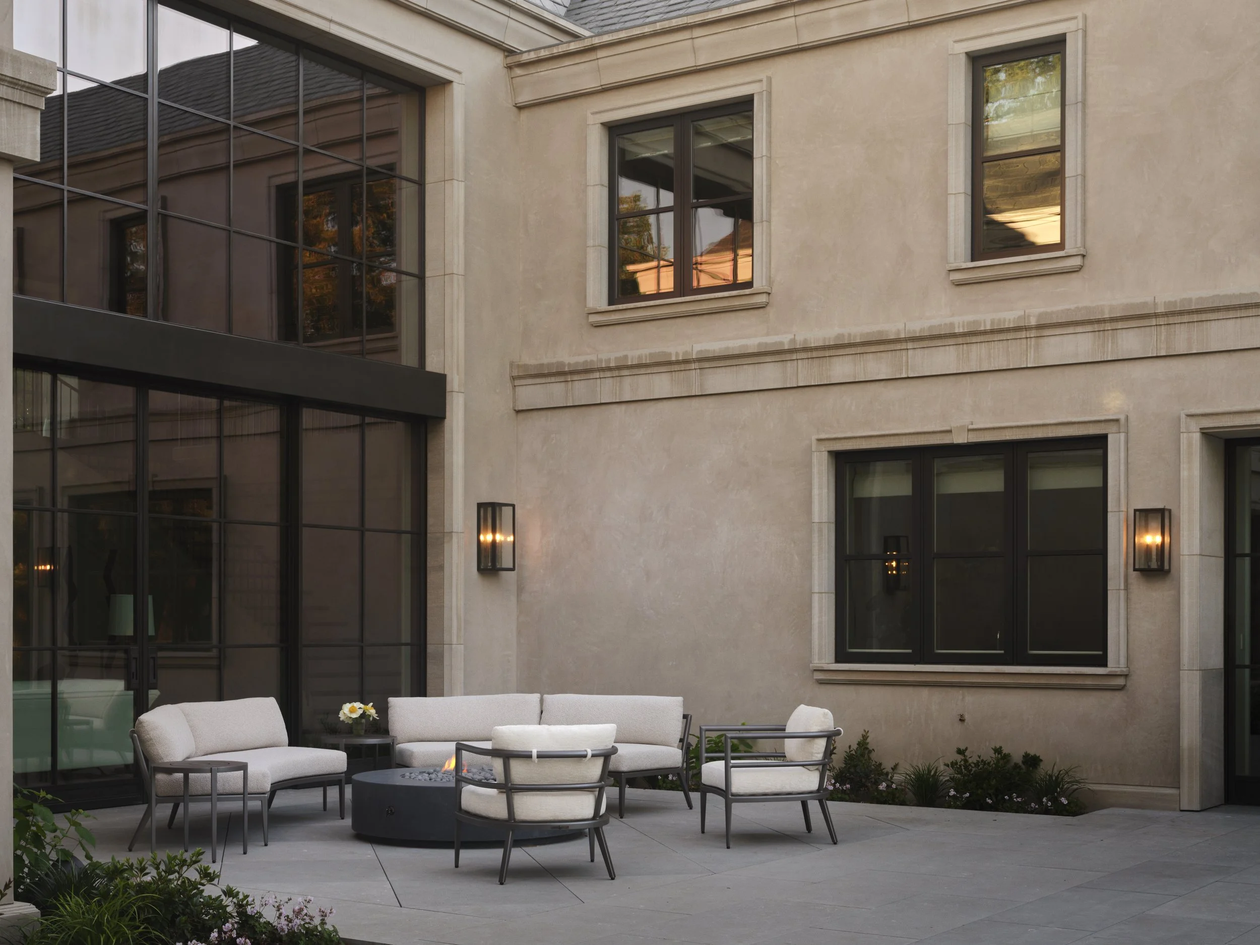 Outdoor patio with beige cushioned seating around a fire pit, flowers, and wall-mounted lights, in front of a modern beige building with large windows.