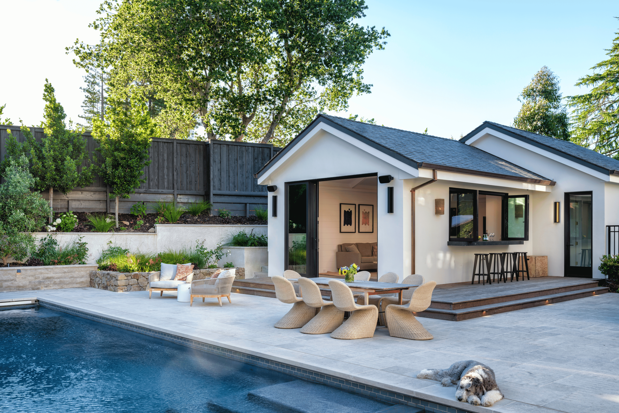 Backyard with swimming pool, outdoor furniture, and a white house with black trim, open sliding door, and wooden deck, surrounded by trees and a wooden fence, with a dog resting on the patio.