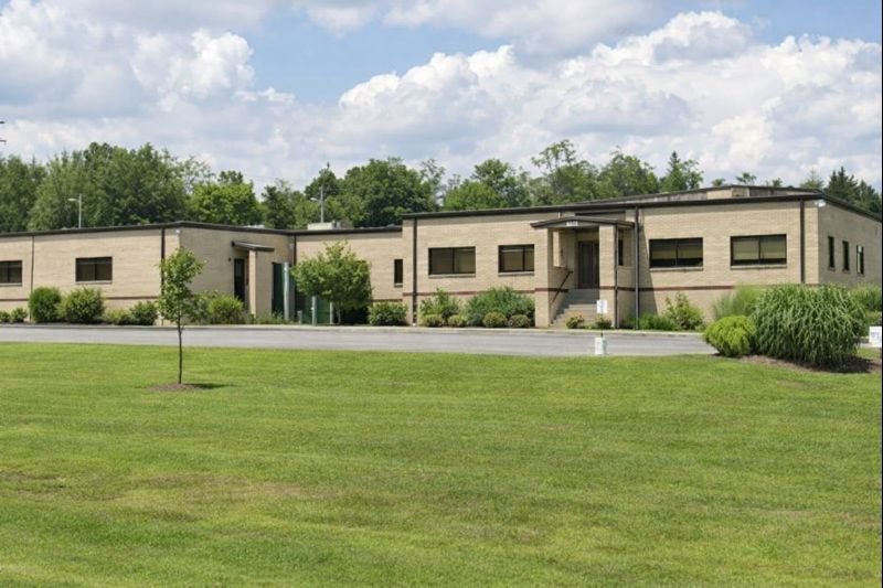 A single-story modern beige building with rectangular windows, surrounded by green grass and small trees, under a partly cloudy sky.