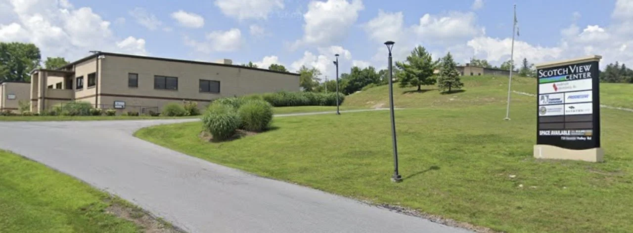 Exterior view of a modern building with a paved driveway, grassy lawn with bushes, and a sign that reads "Scotch View Center" on a sunny day with scattered clouds.