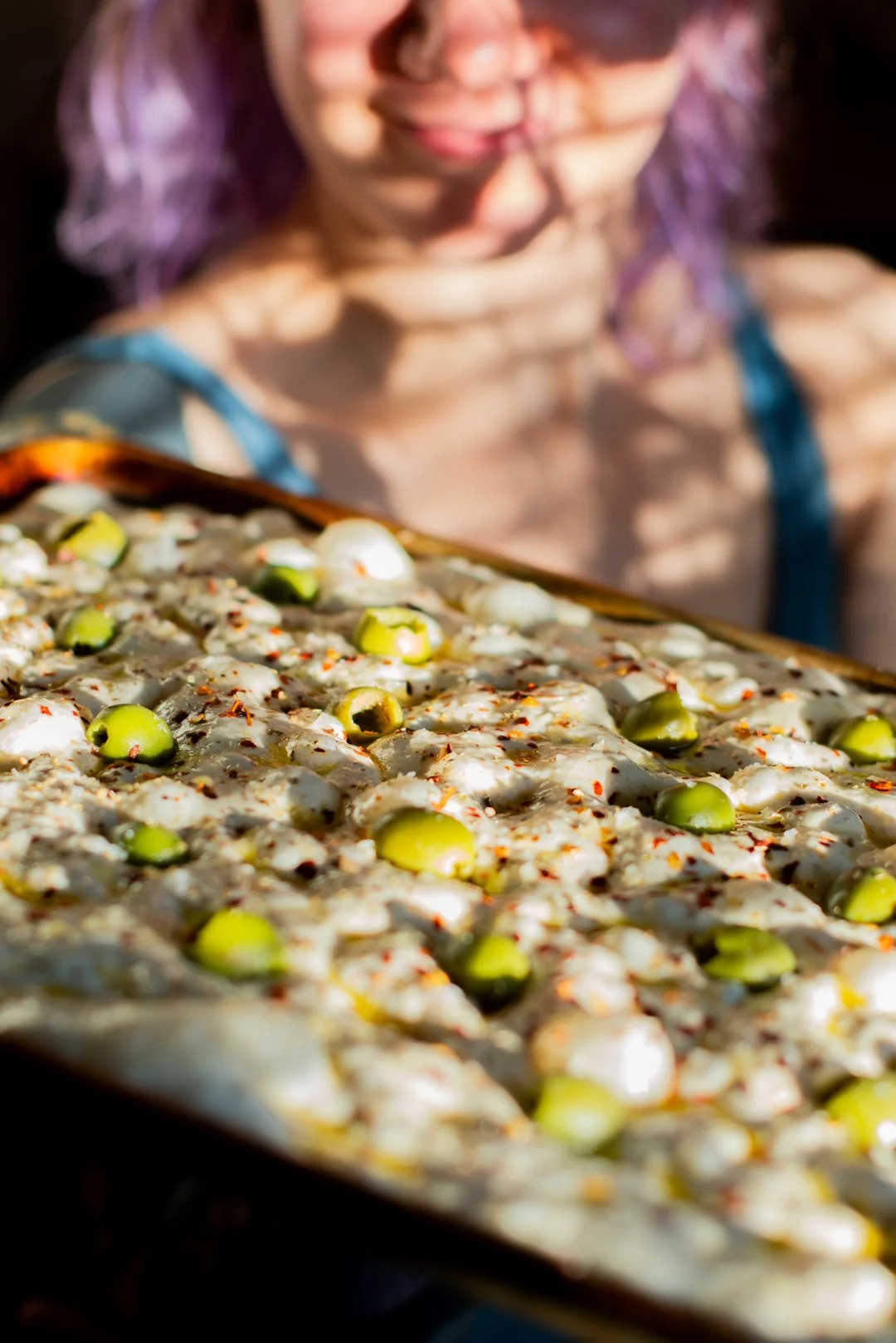 A person with purple hair looks down at a large tray of unbaked focaccia dough topped with peppers or olives, garnished with red chili flakes, outdoors in sunlight.