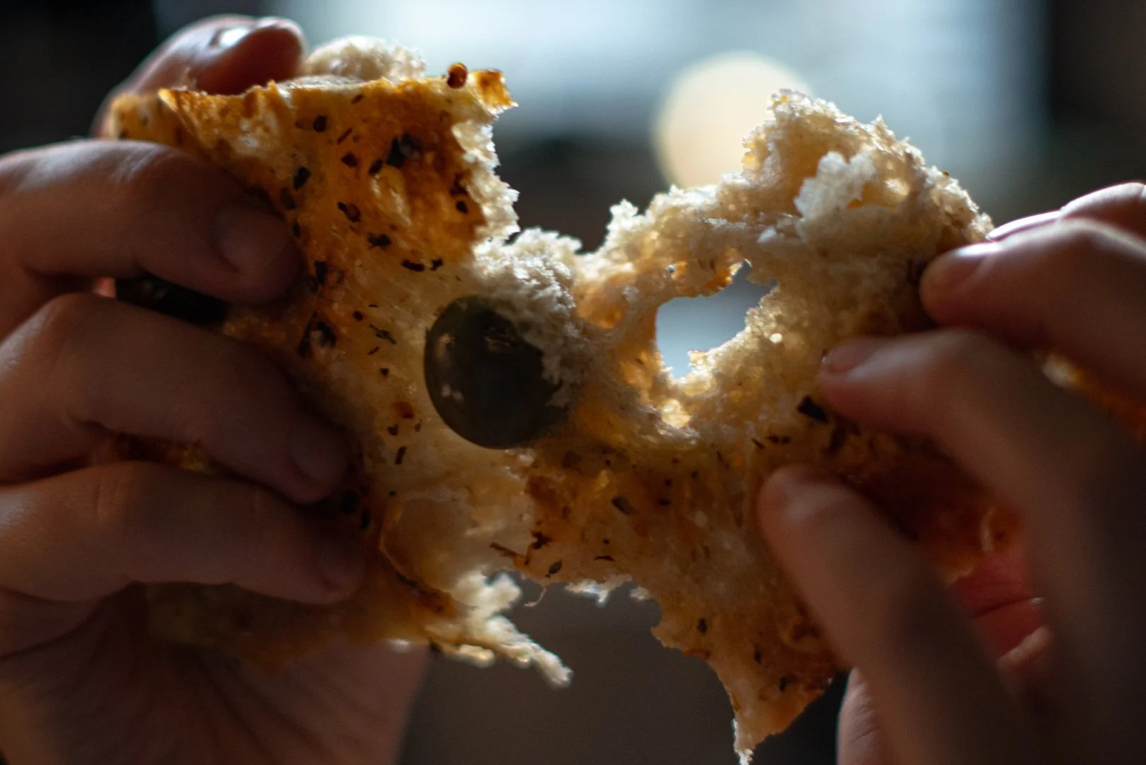 Two hands tearing apart a piece of thin, crispy, focaccia bread with green Castlevetrano olives, revealing its soft interior.