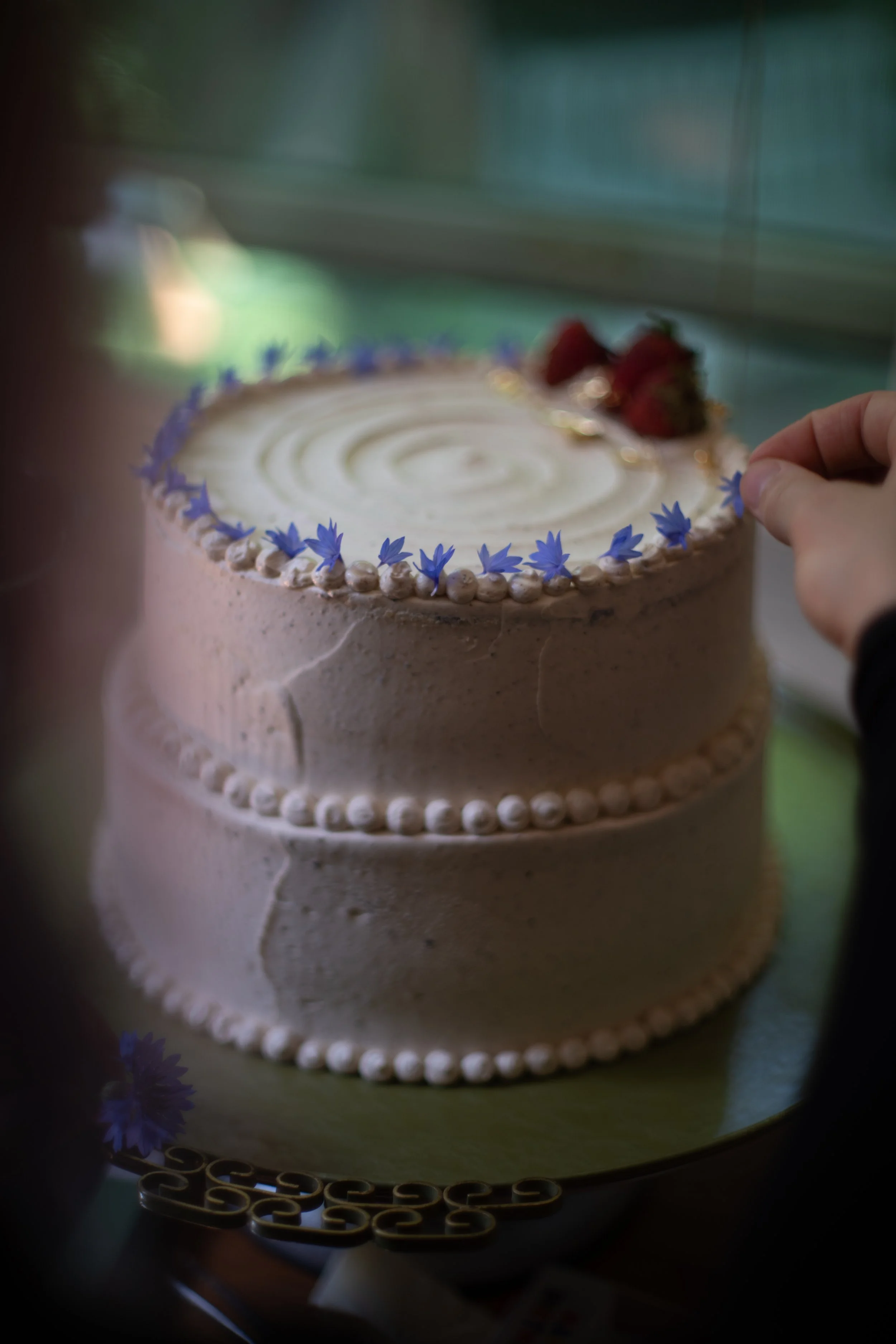 Close-up of a two-tiered birthday cake with white frosting, decorated with small purple flowers around the edge, with strawberries on top, being touched by a person's hand.