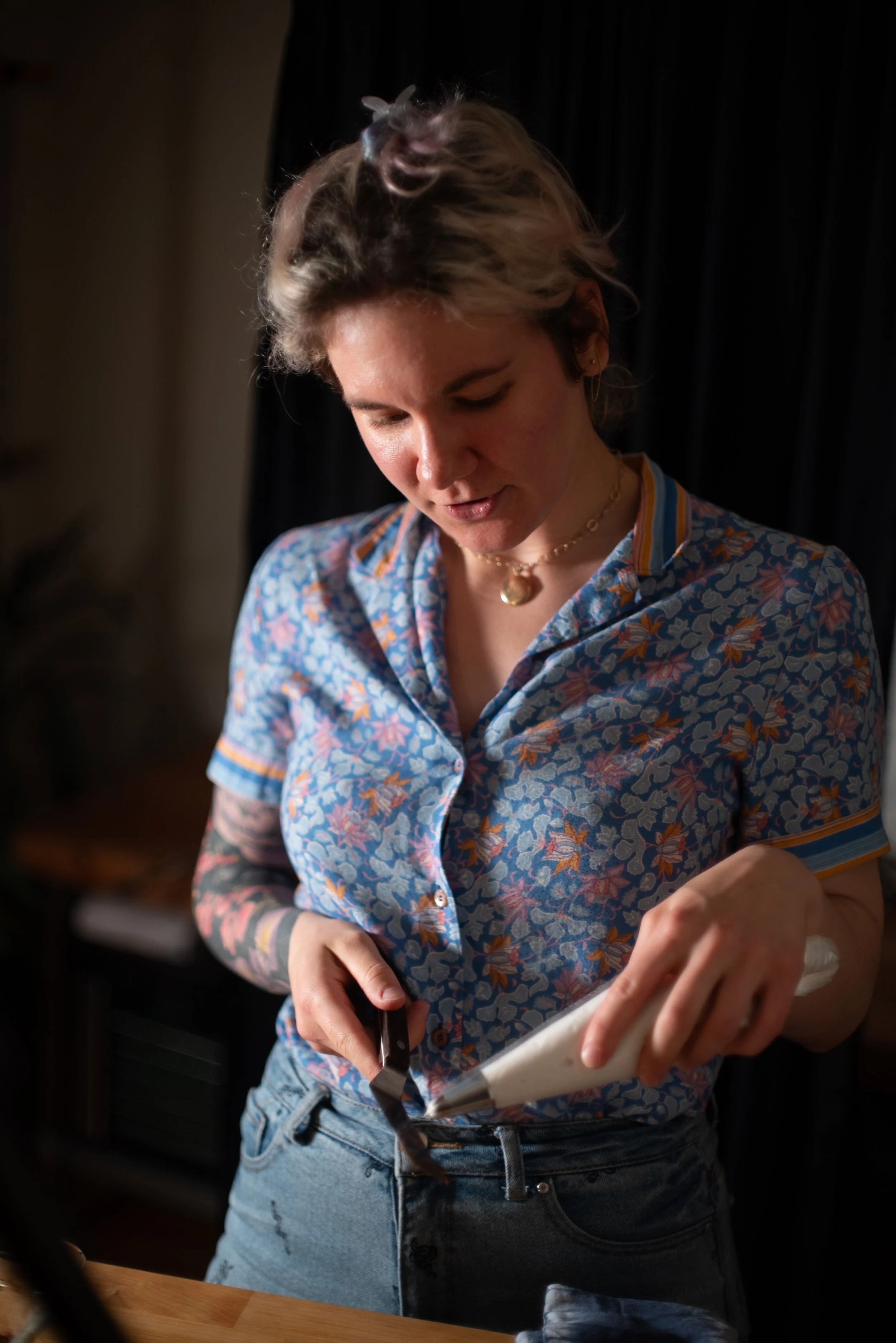 A woman with short, light-colored hair is standing in a dimly lit room, wearing a blue floral shirt and jewelry, while spreading chocolate frosting onto a cupcake with a piping bag.