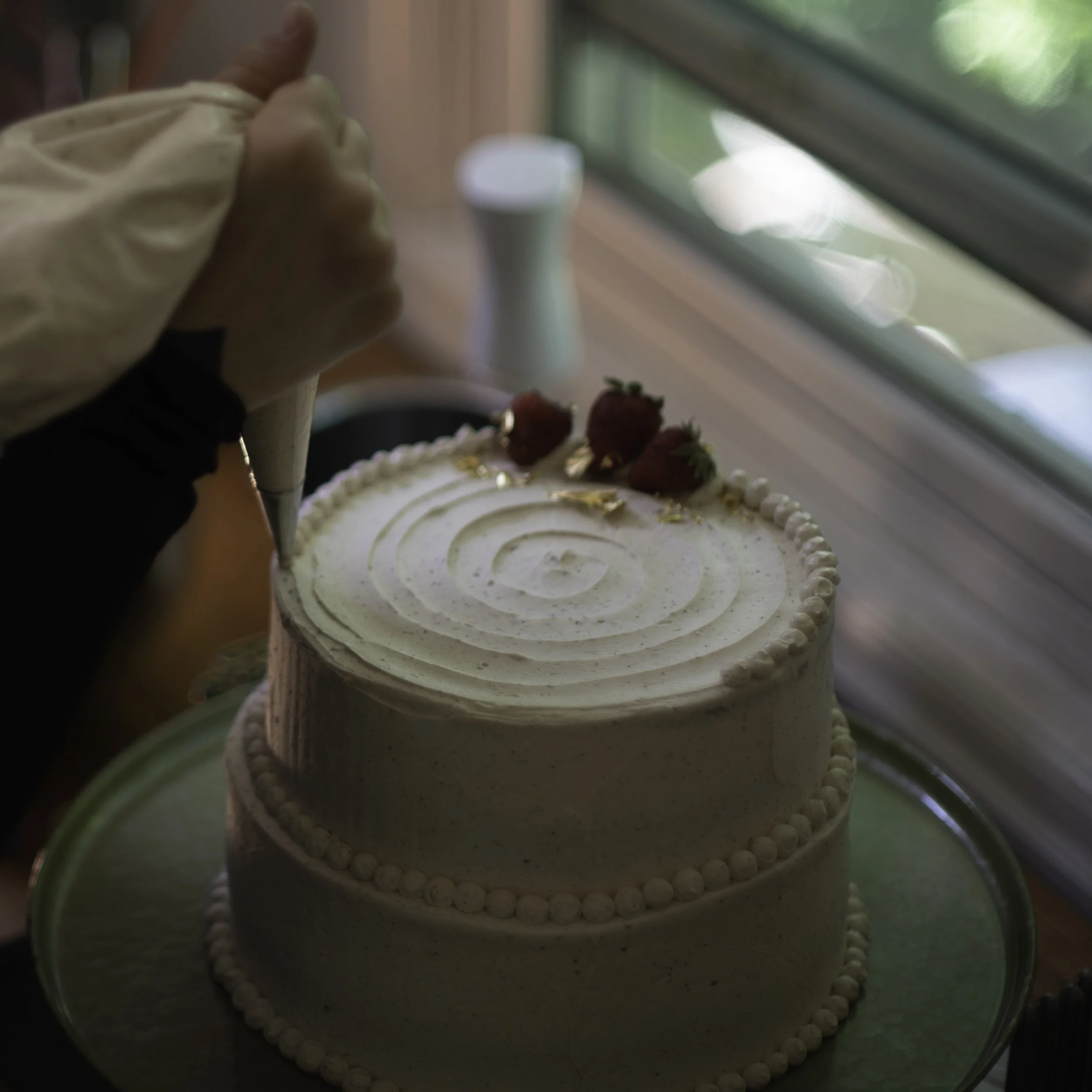 Person piping white frosting onto a two-layer cake decorated with strawberries.