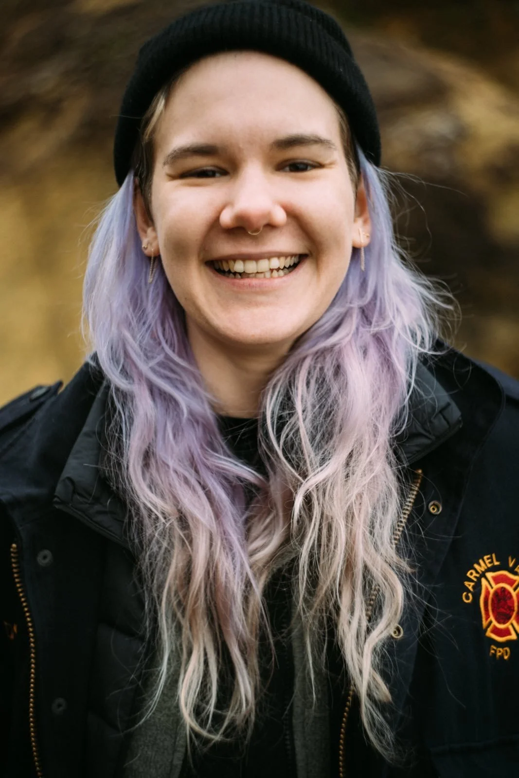 A young woman with long, wavy lavender hair, smiling outdoors, wearing a black beanie and a dark jacket with a fire department patch.