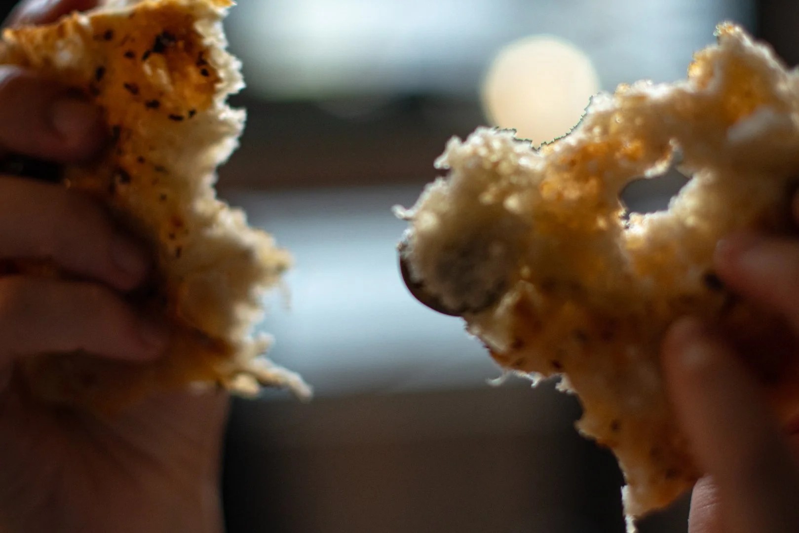 Two hands holding torn pieces of focaccia bread with green Castlevetrano olives, against a blurred background with light source.