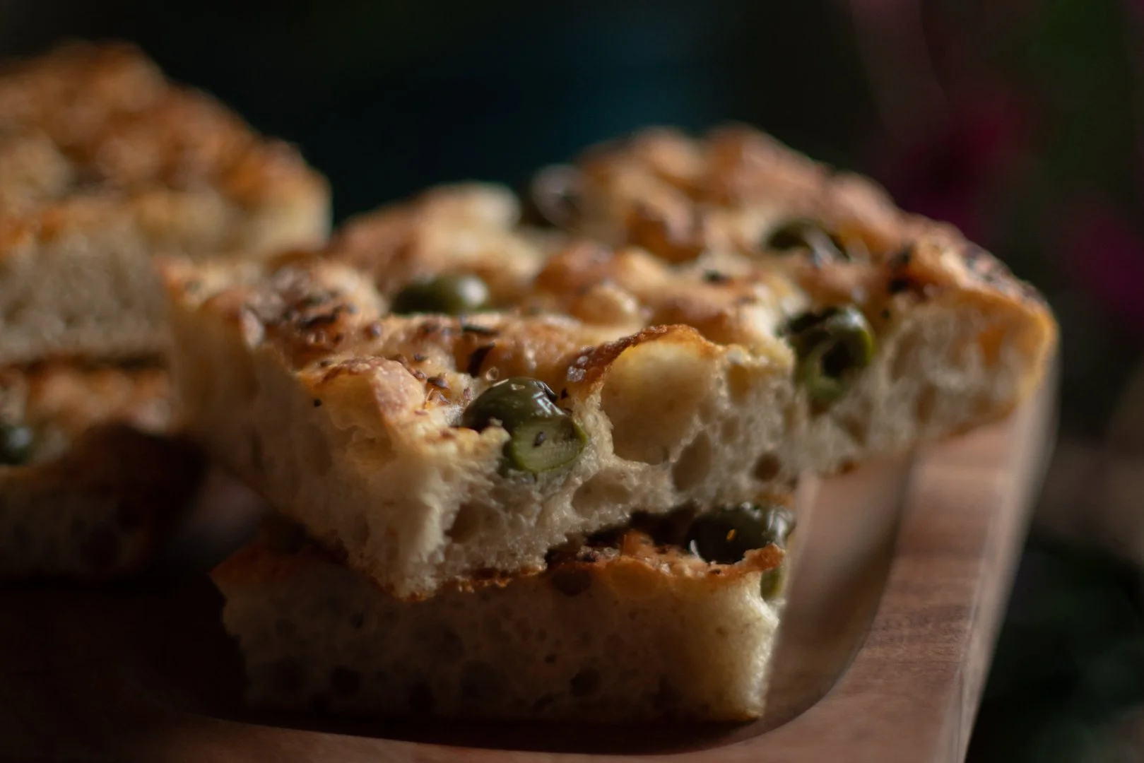 Close-up of a slice of bread with green Castlevetrano olives, flakey salt, and herbs on top, resting on a wooden surface.