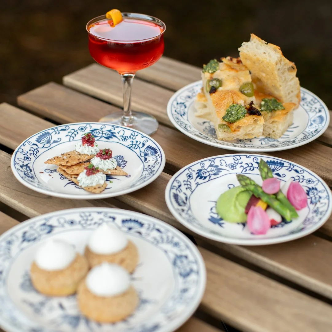 Assorted appetizers and desserts on blue and white floral plates, including a red cocktail with a lemon twist, focaccia, crackers with cheese and toppings, a colorful salad with radishes, and cream-topped pastries, all on a wooden outdoor table.