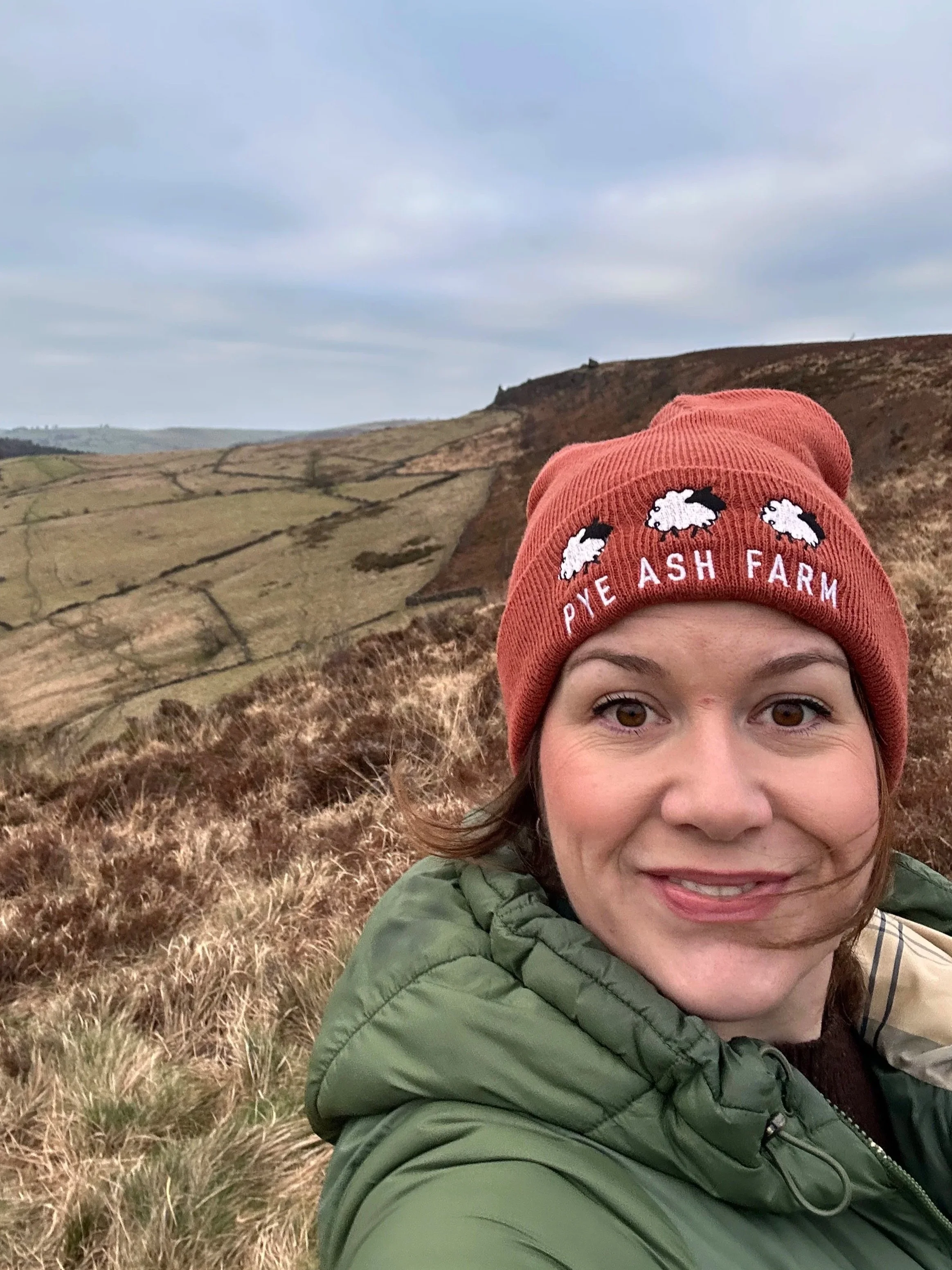 A woman taking a selfie outdoors on a cloudy day, wearing a red beanie with embroidered sheep and the words 'Pye Ash Farm', and a green jacket. She is standing on a hillside with grassy and brown terrain, with rolling green hills and a cloudy sky in the background.
