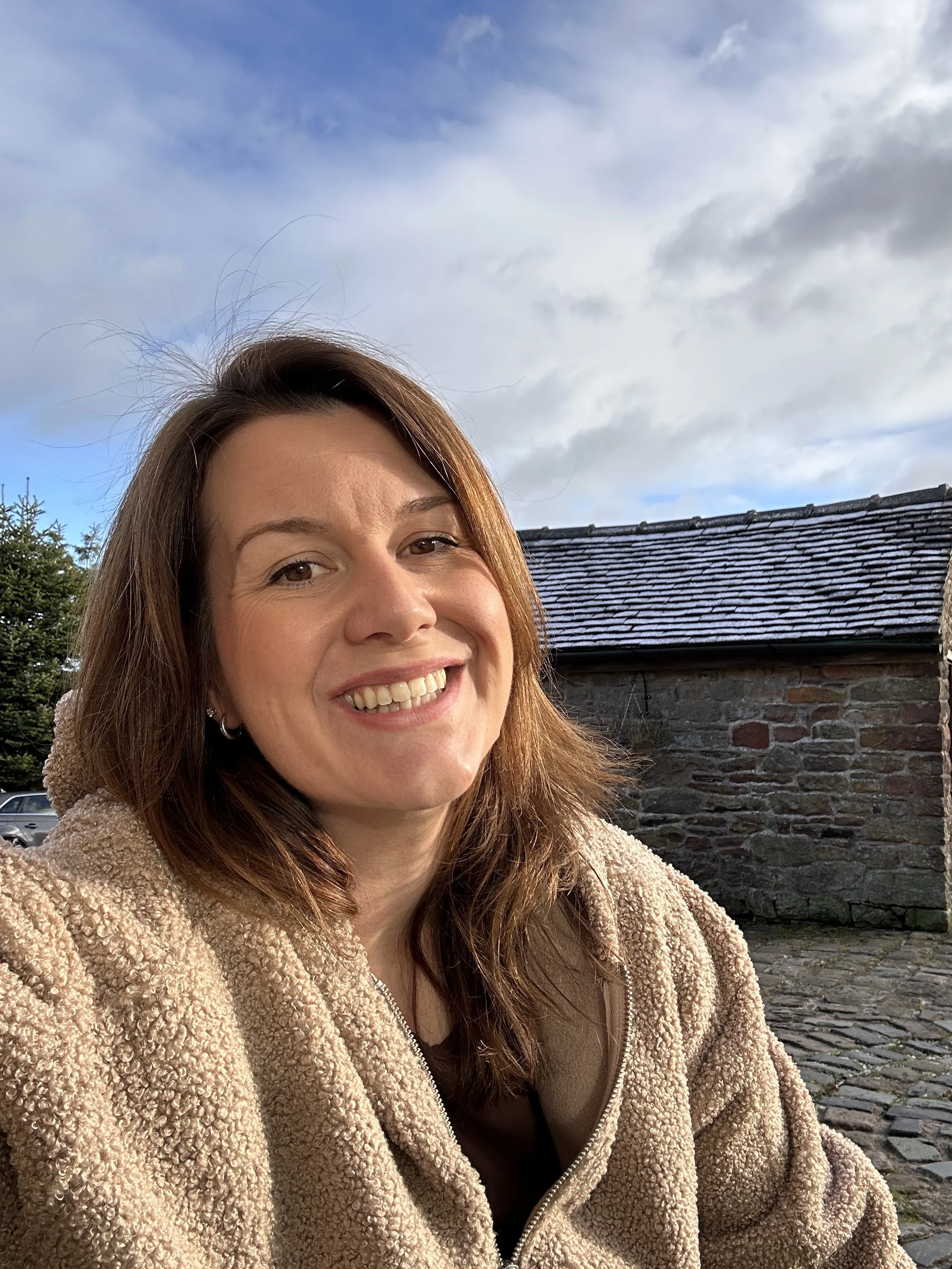 A woman with shoulder-length brown hair smiling outdoors against a cloudy sky and a stone building with a tiled roof.