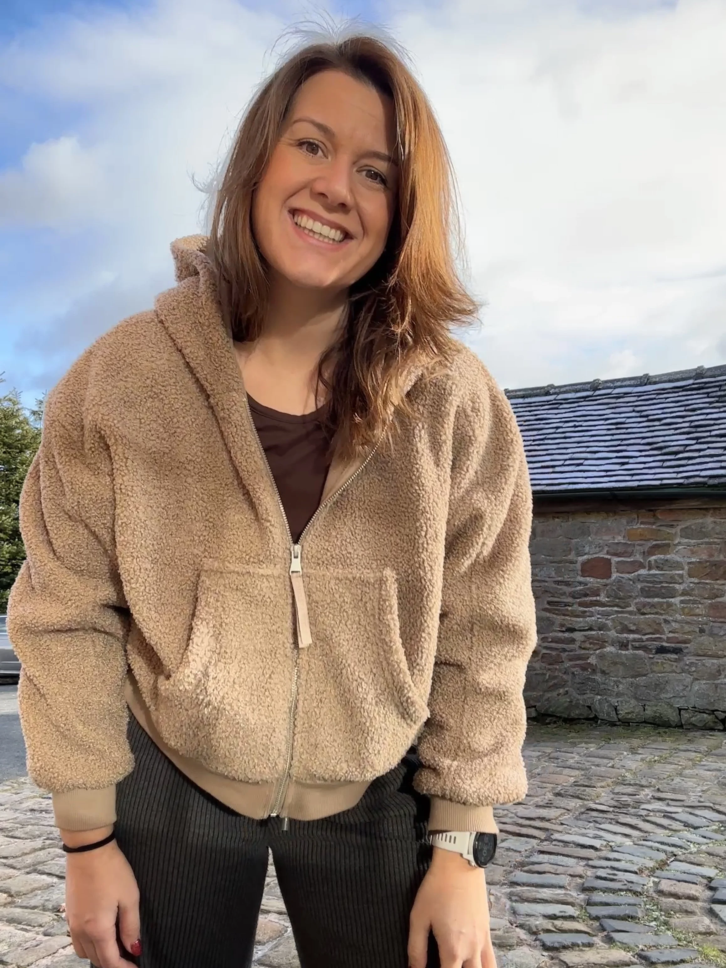 A young woman with shoulder-length red hair smiling outdoors wearing a tan fleece jacket and black pants, standing on a cobblestone street with a stone building in the background.