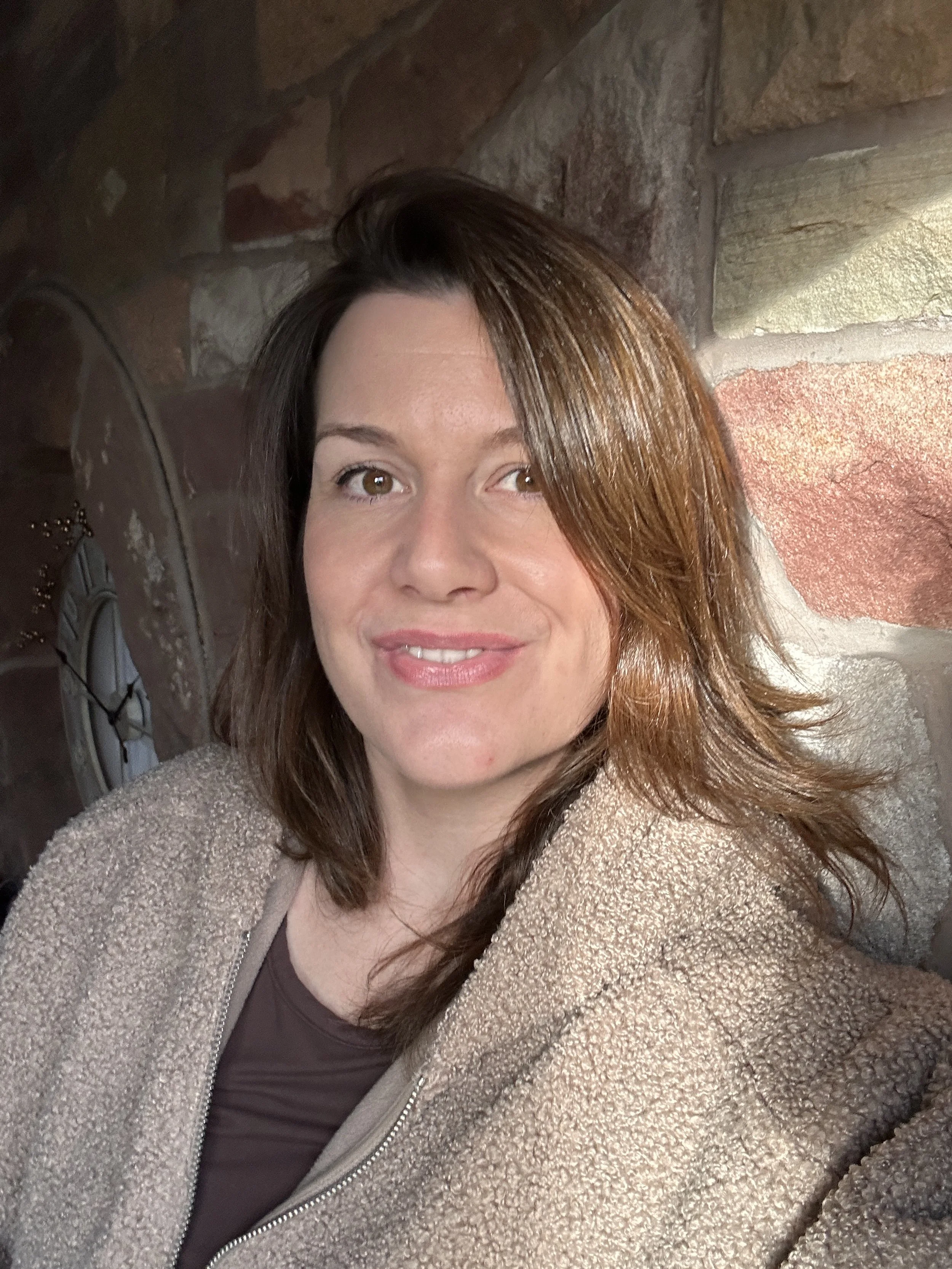 A woman with shoulder-length brown hair smiling, wearing a beige fleece jacket, in front of a stone wall and a clock.