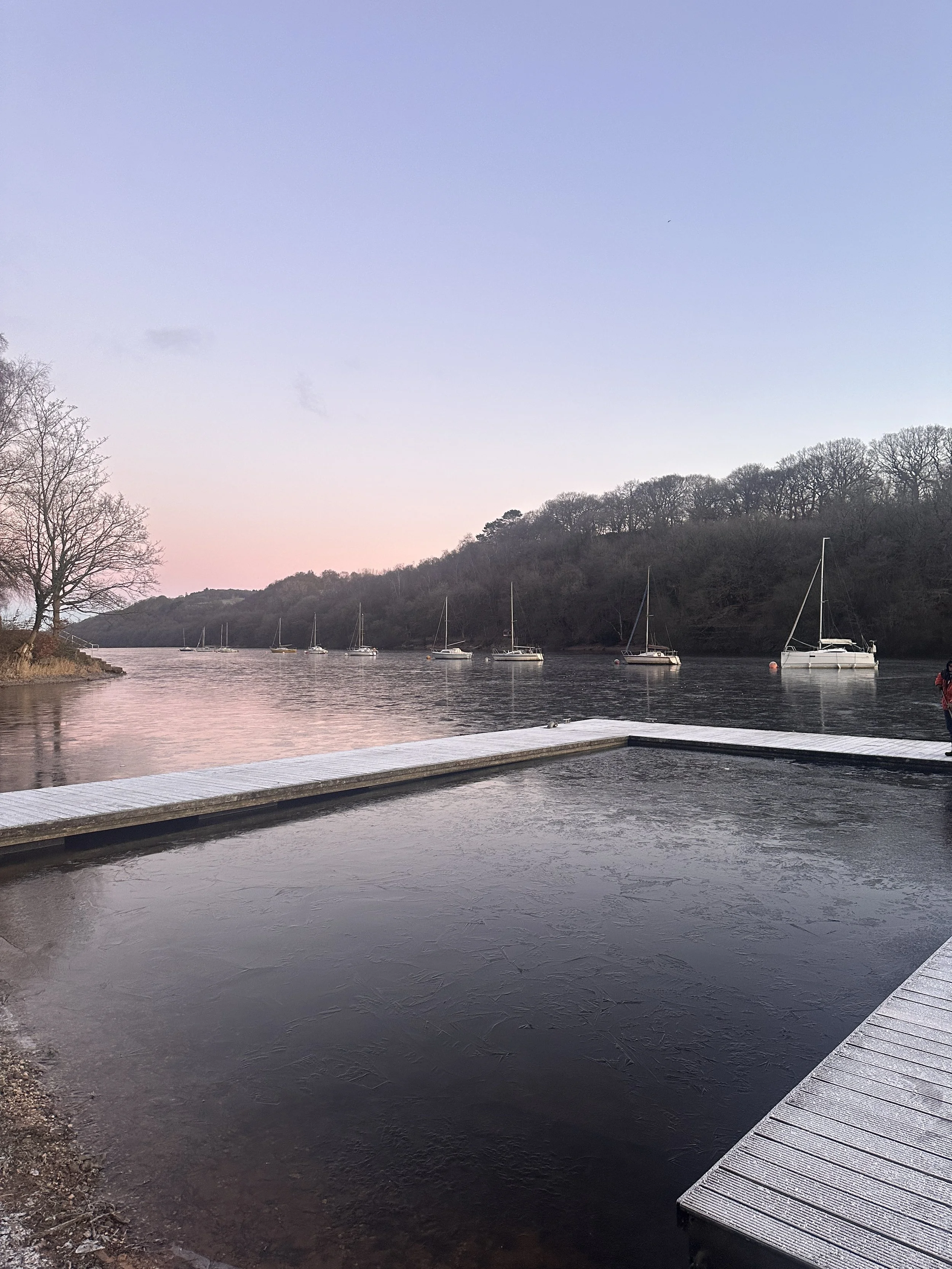 A serene, calm, trusting lakeside scene at dusk with sailboats anchored on calm water and a snow or ice-covered dock in foreground. Bare trees and hills are visible in the distance against a pastel sky.