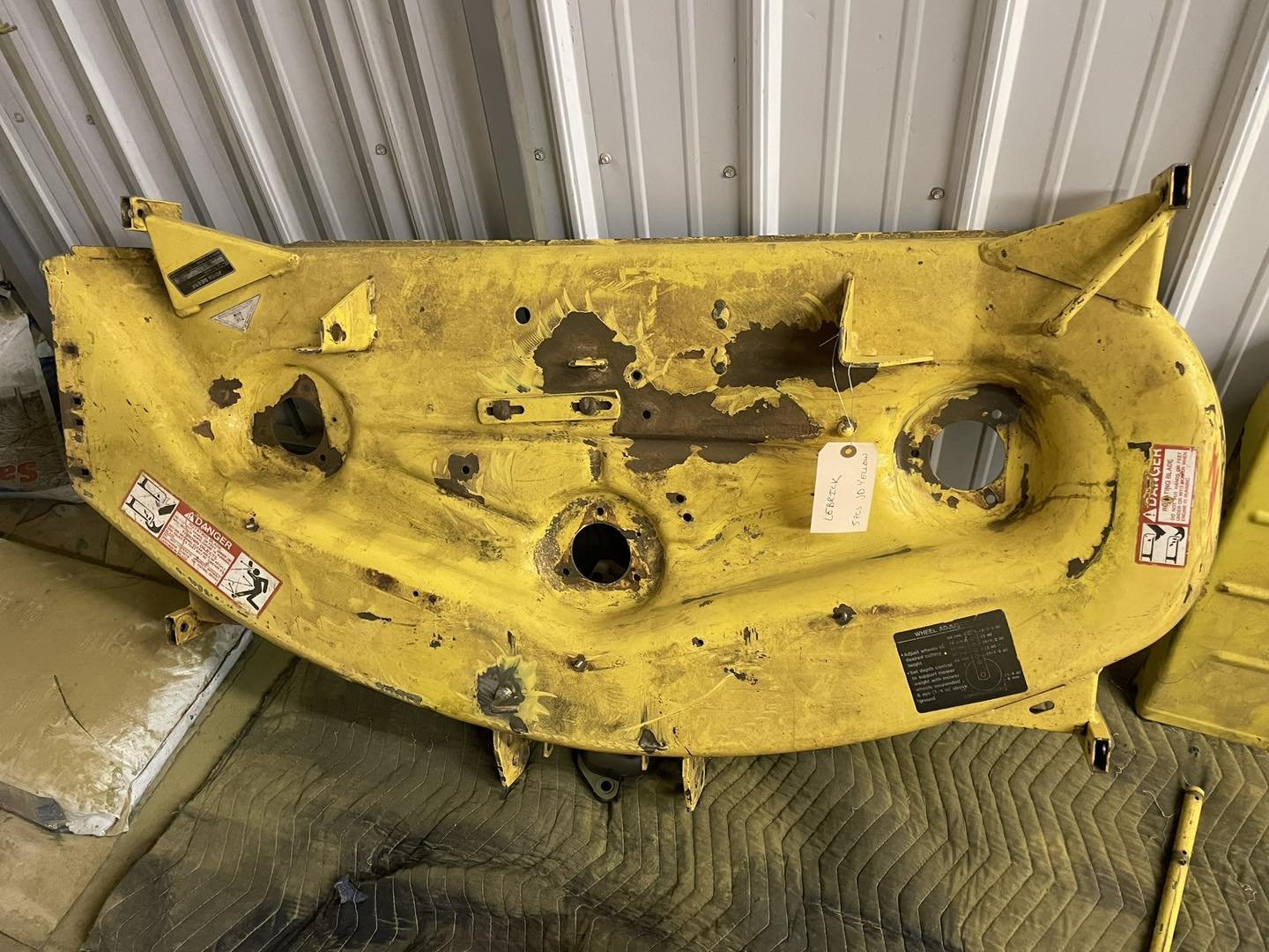 Yellow tractor attachment with scratches, holes, and warning labels, placed on a padded surface in front of a metal wall.