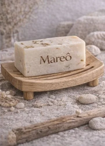 Sabão artesanal com a palavra "Maré" escrito, colocado sobre um suporte de madeira sobre uma superfície de pedra com pedras ao redor.
