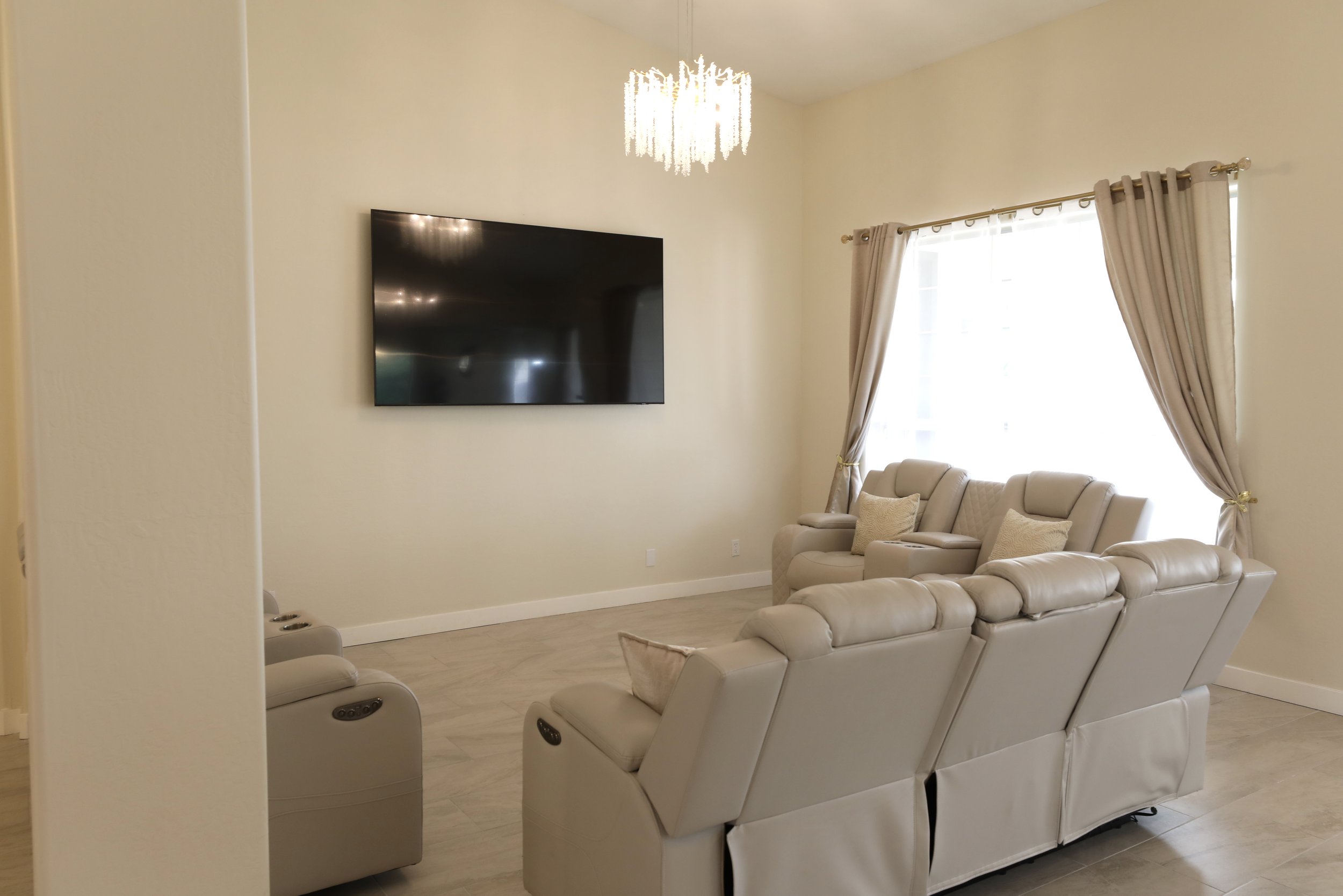 A living room with a wall-mounted flat-screen TV, beige leather recliner sofa, armchair with pillow, large window with beige curtains, and a modern chandelier ceiling light.