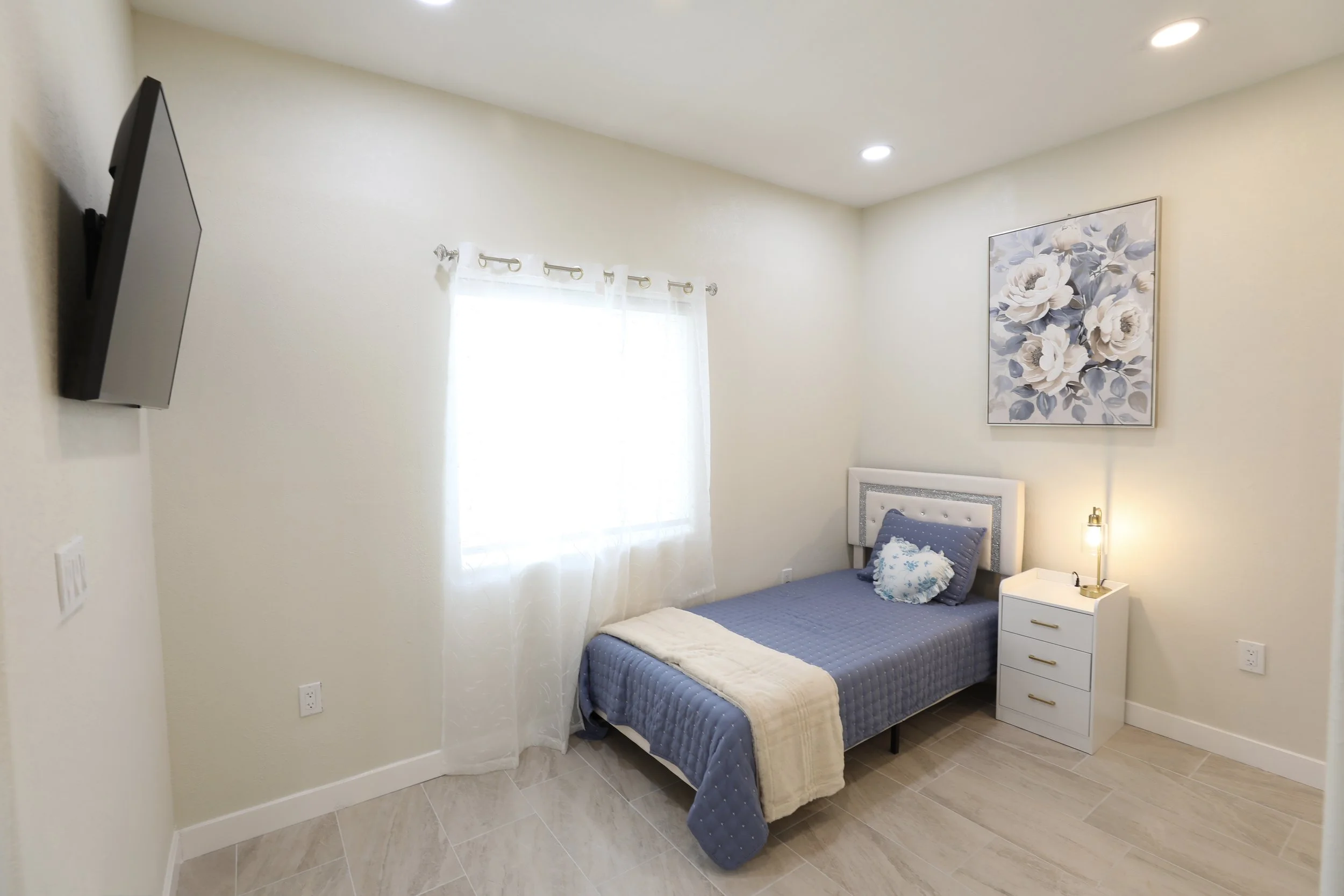 Small bedroom with a single bed, blue quilt, white headboard, pillows, a nightstand with a lamp, floral wall art, window with curtains, and a wall-mounted TV.