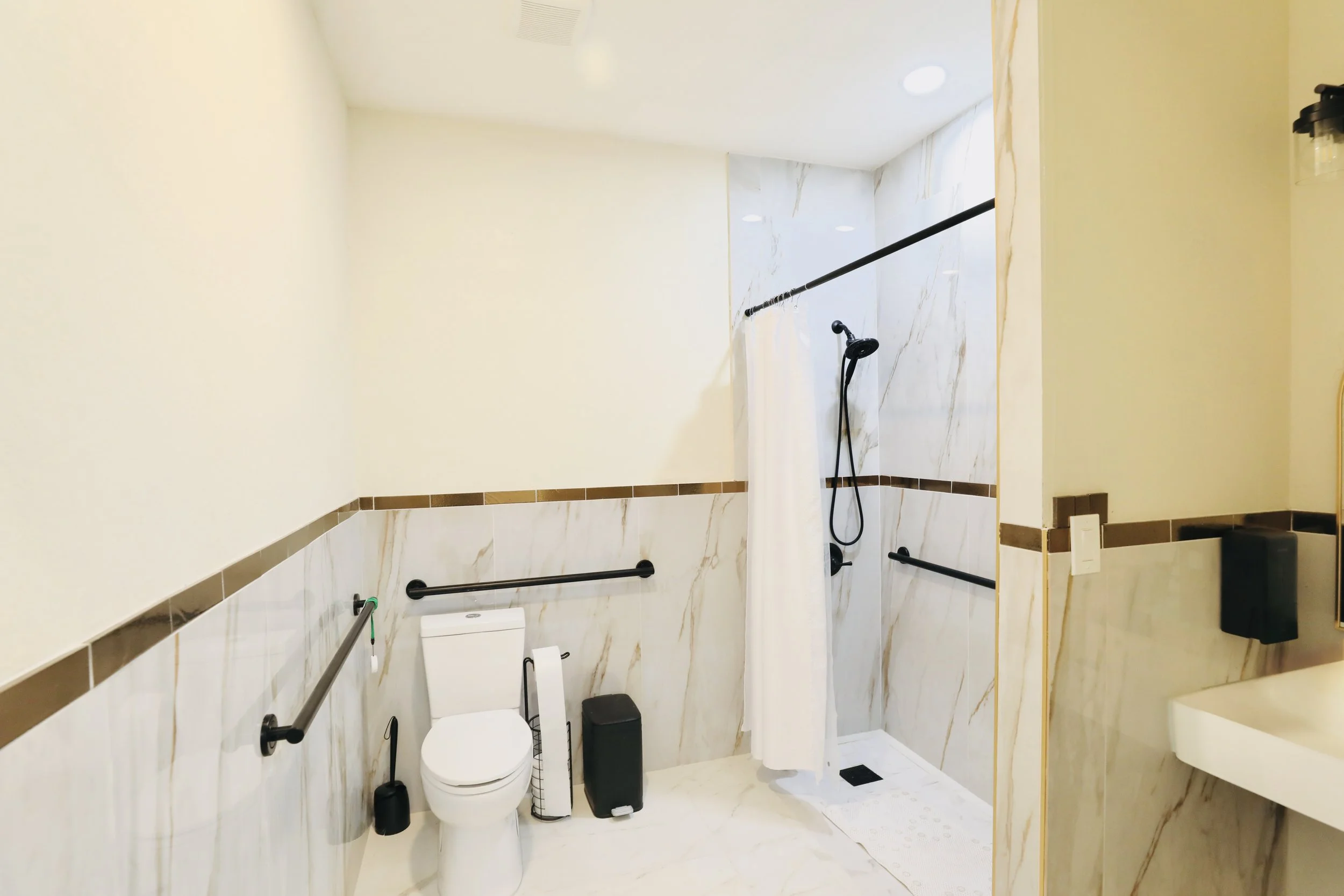 A bathroom with a walk-in shower, toilet, and vanity with a sink. The shower has a curtain, black shower head, and grab bars. The bathroom has beige walls with marble tiles and brown trim.
