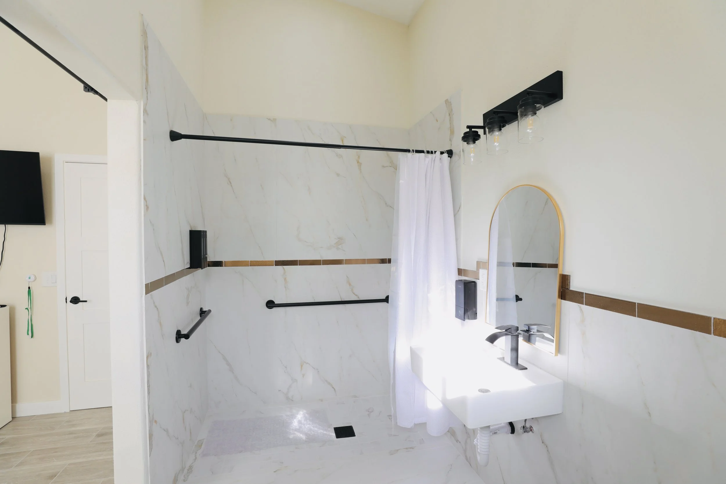 Accessible bathroom with marble walls, black grab bars, white shower curtain, small sink, mirror, and modern lighting.
