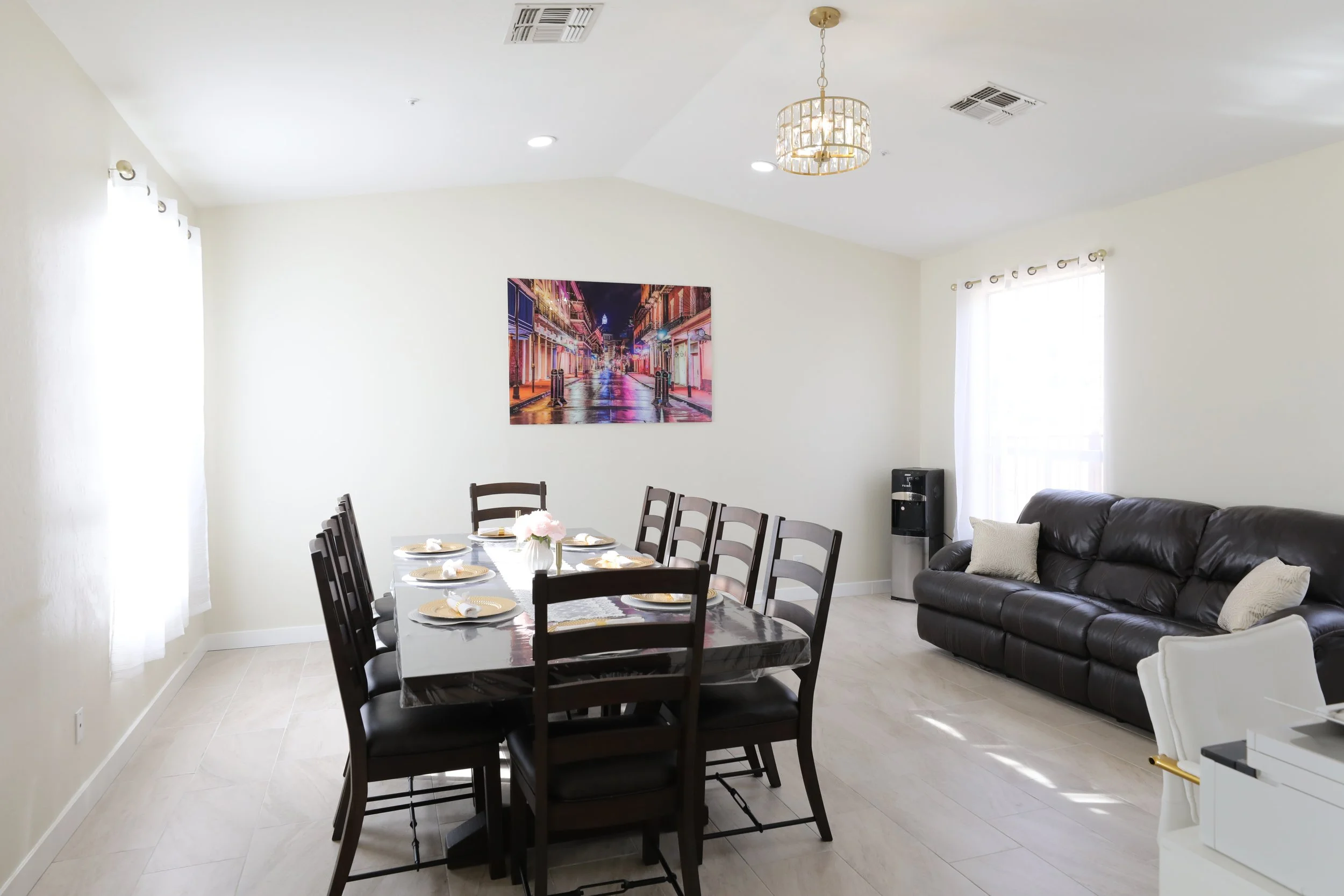 Dining room with a wooden dining table set for six, a dark leather sofa with throw pillows, a water dispenser, a window with white curtains, a framed colorful city street photograph, and a chandelier hanging from the ceiling.