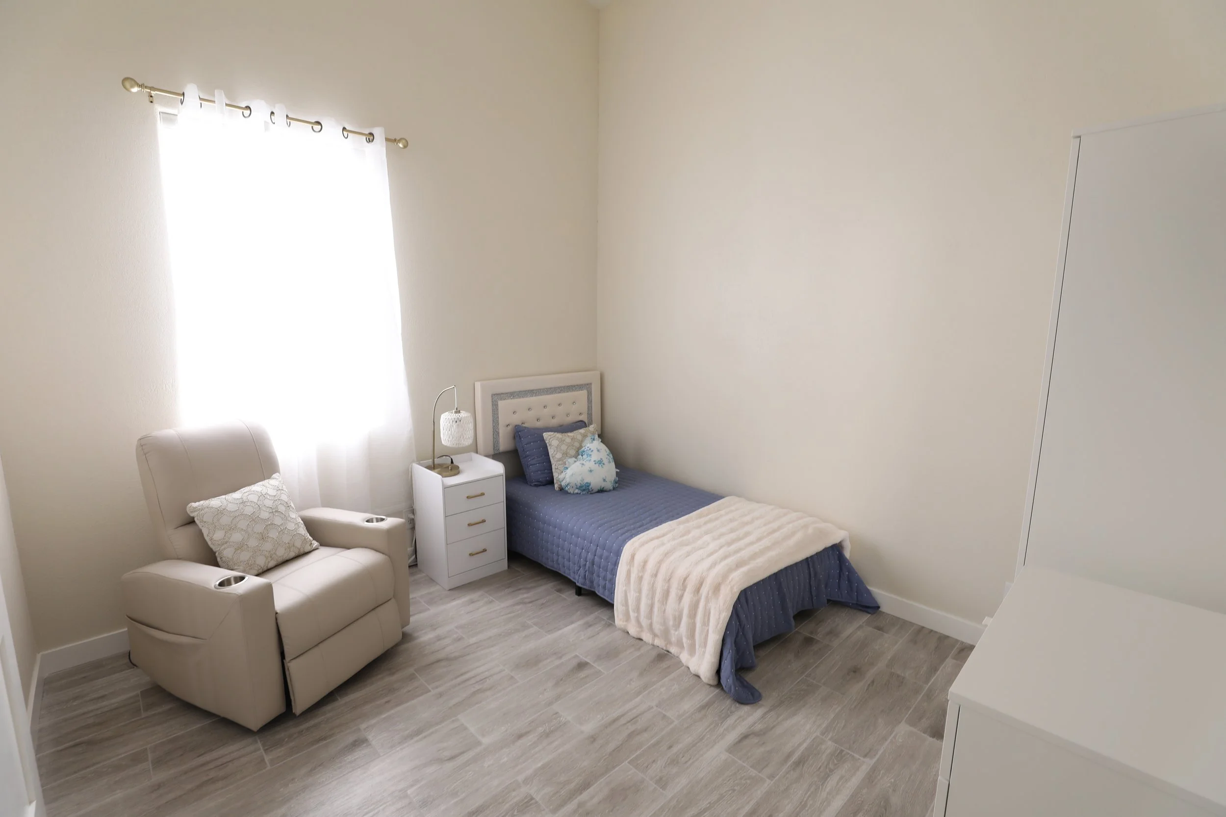 A small bedroom with a single bed with blue bedding and decorative pillows, a white nightstand with a lamp, a beige recliner chair with a cushion, a white dresser, and a window with white curtains allowing sunlight to brighten the room.
