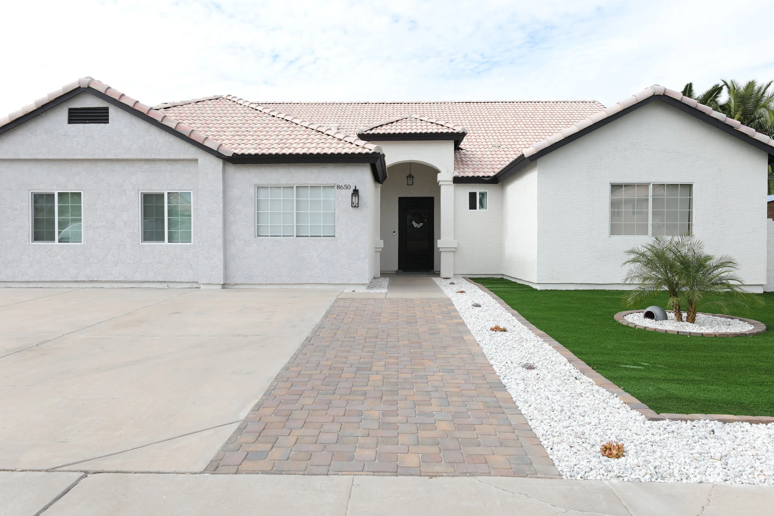 Front view of a modern house with a brick pathway leading to the black front door, a small landscaped yard with green grass, a palm tree, and decorative white stones.