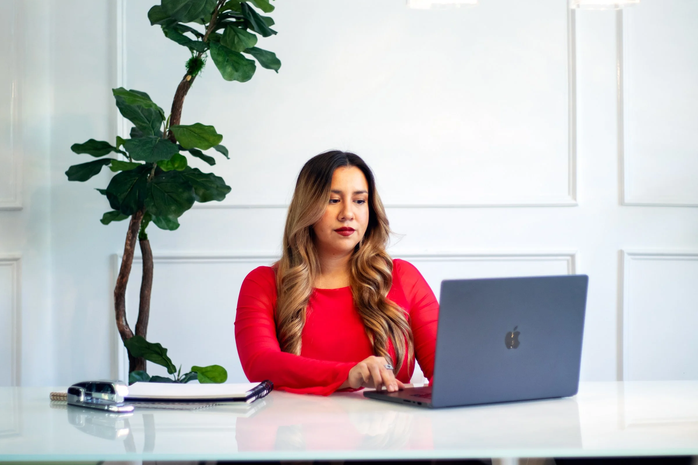 A woman with long wavy hair wearing a red top is sitting at a white desk working on a silver laptop. There is a large green indoor plant behind her on the left side and a notebook with a pen and some small objects on the desk.