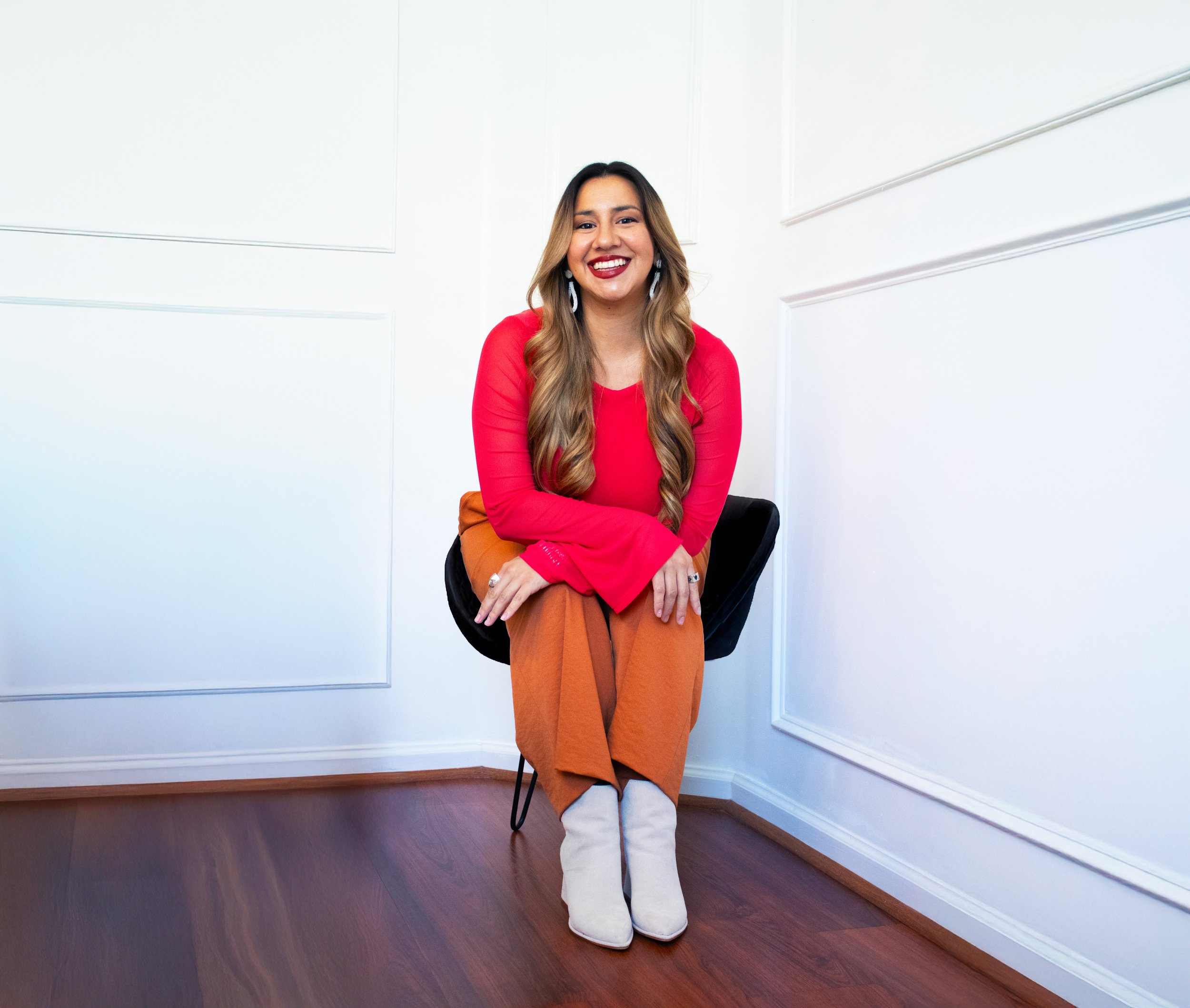 A woman with long wavy hair, wearing a red top, brown pants, white boots, and earrings, sitting on a black chair in a white room with wooden flooring, smiling at the camera.