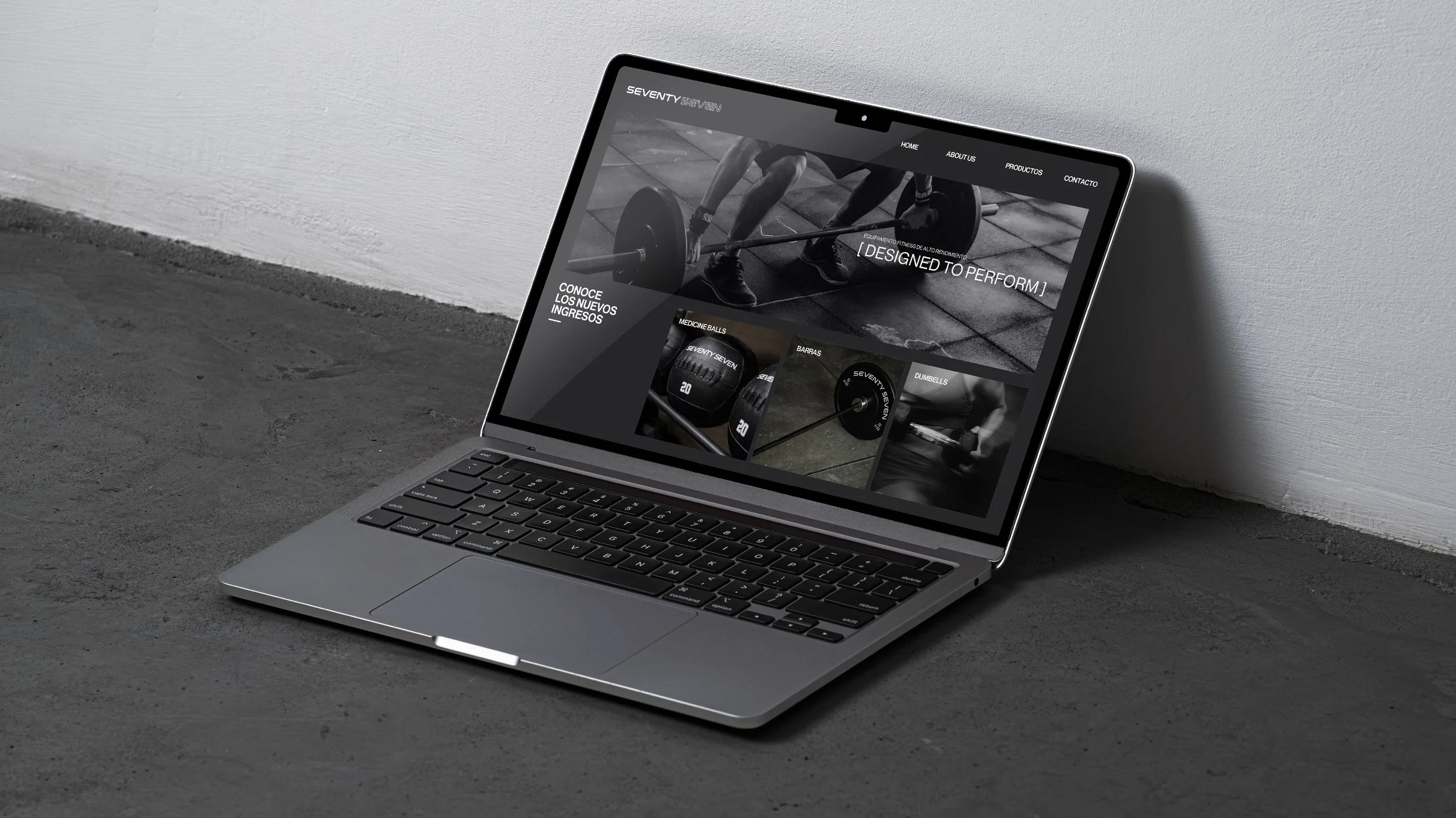 Portátil gris con pantalla mostrando un sitio web de equipo para entrenamiento de fuerza, incluyendo balones medicinales, barras y mancuernas, sobre un suelo de concreto junto a una pared blanca.