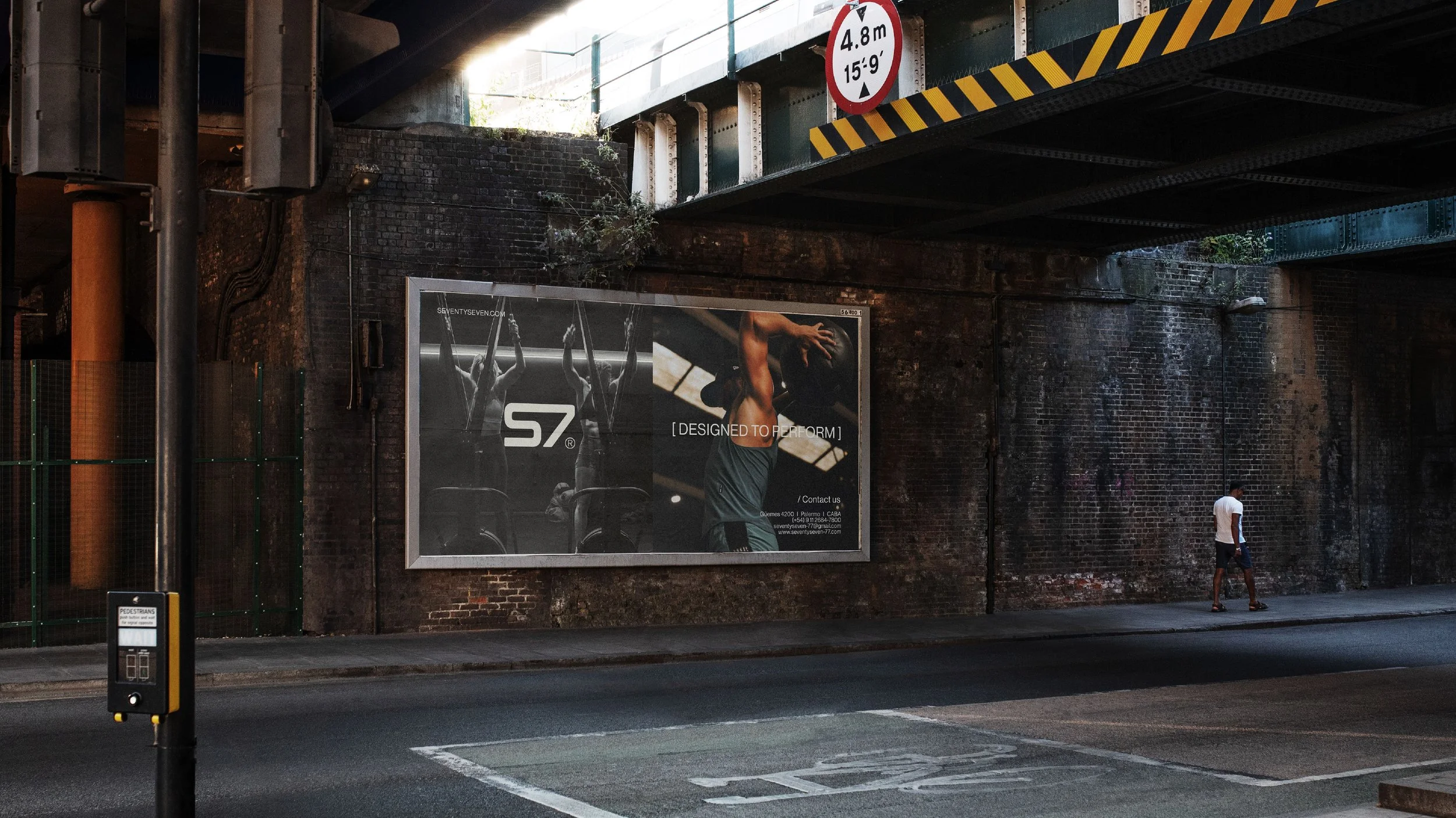 Un cartel publicitario en una pared de ladrillos bajo un puente que muestra una imagen de una mujer haciendo ejercicio, con un texto que dice 'Designed to Perform'. En la esquina superior derecha, un signo de tráfico que indica una altura máxima de 4.8 metros o 15 pies y 9 pulgadas.