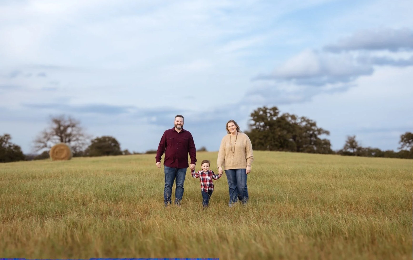 A family of three walking hand in hand through a grassy field, with trees and hay bales in the distance and a cloudy sky overhead.