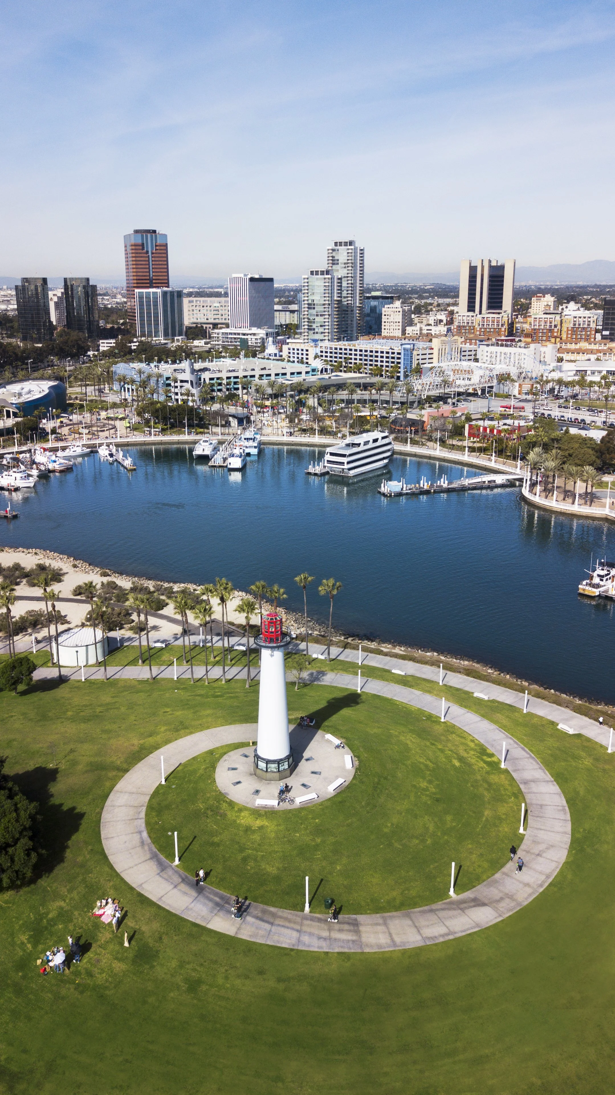 Aerial view of a cityscape featuring a marina with boats, a lighthouse on a grassy park, and tall modern buildings in the background under a clear blue sky.