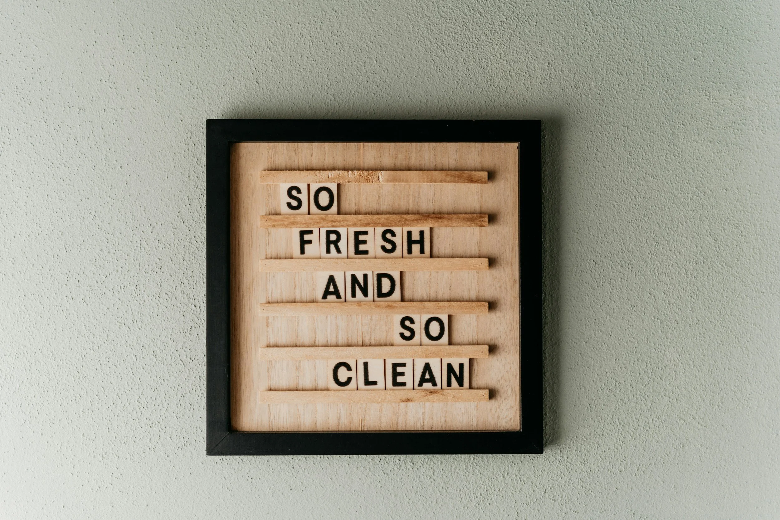 A framed board with a bamboo background displaying the message 'SO FRESH AND SO CLEAN' spelled out with black letter tiles on horizontal slats.