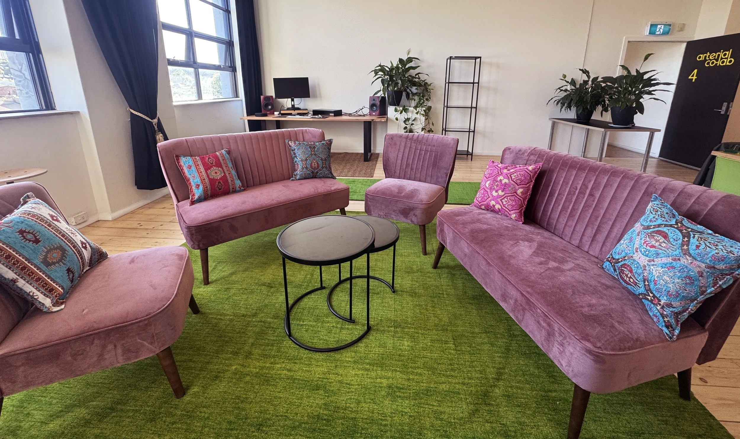 A cozy lounge area with pink velvet sofas and armchairs arranged around a small black round coffee table on a green rug. There are decorative pillows on the sofas, and a desk with a computer and speakers in the background, along with some potted plan
