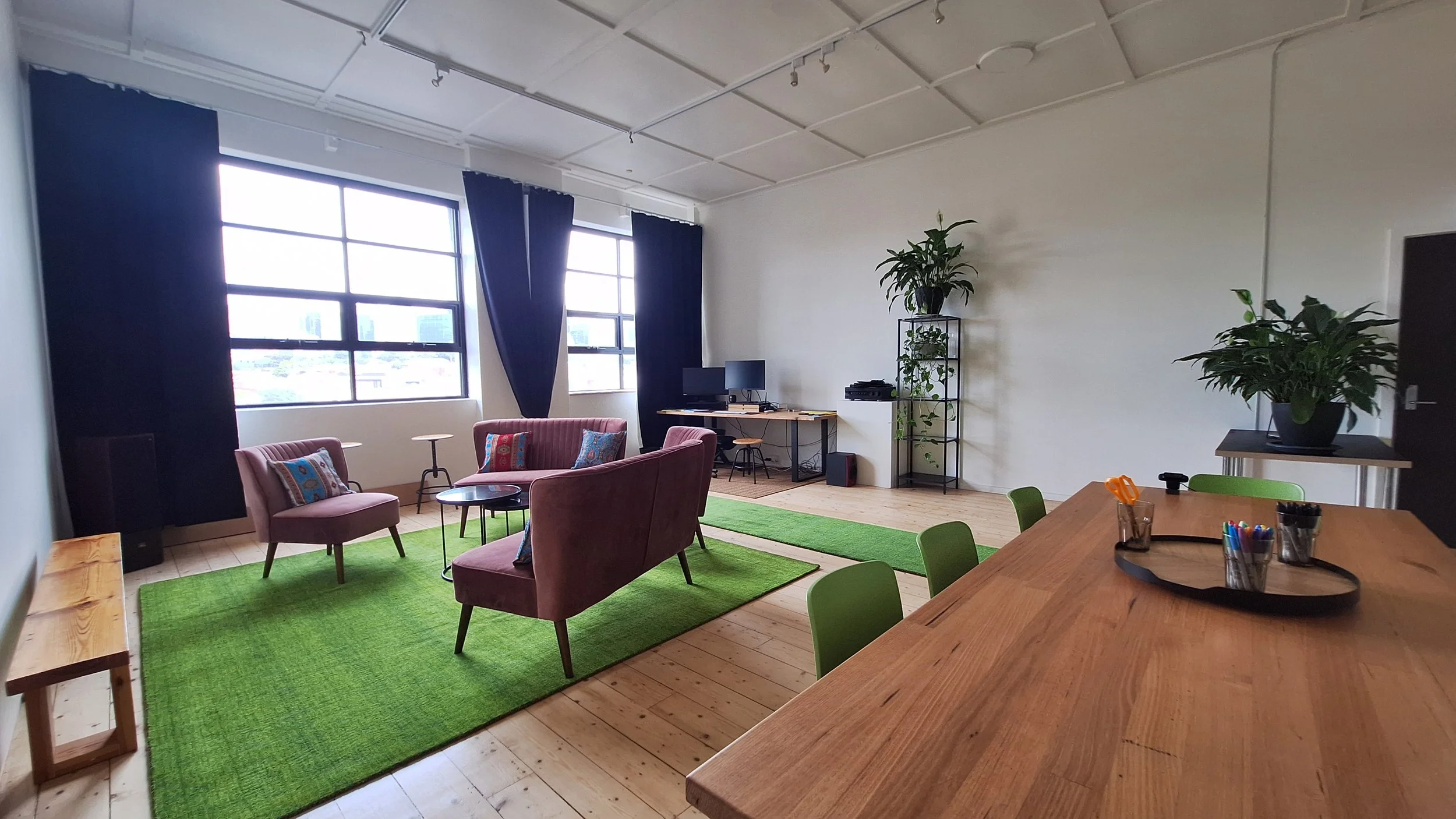 spacious studio space with large windows and wooden floor. The space is filled with a large oak conference table, set of couches and armchairs, and computer table setup