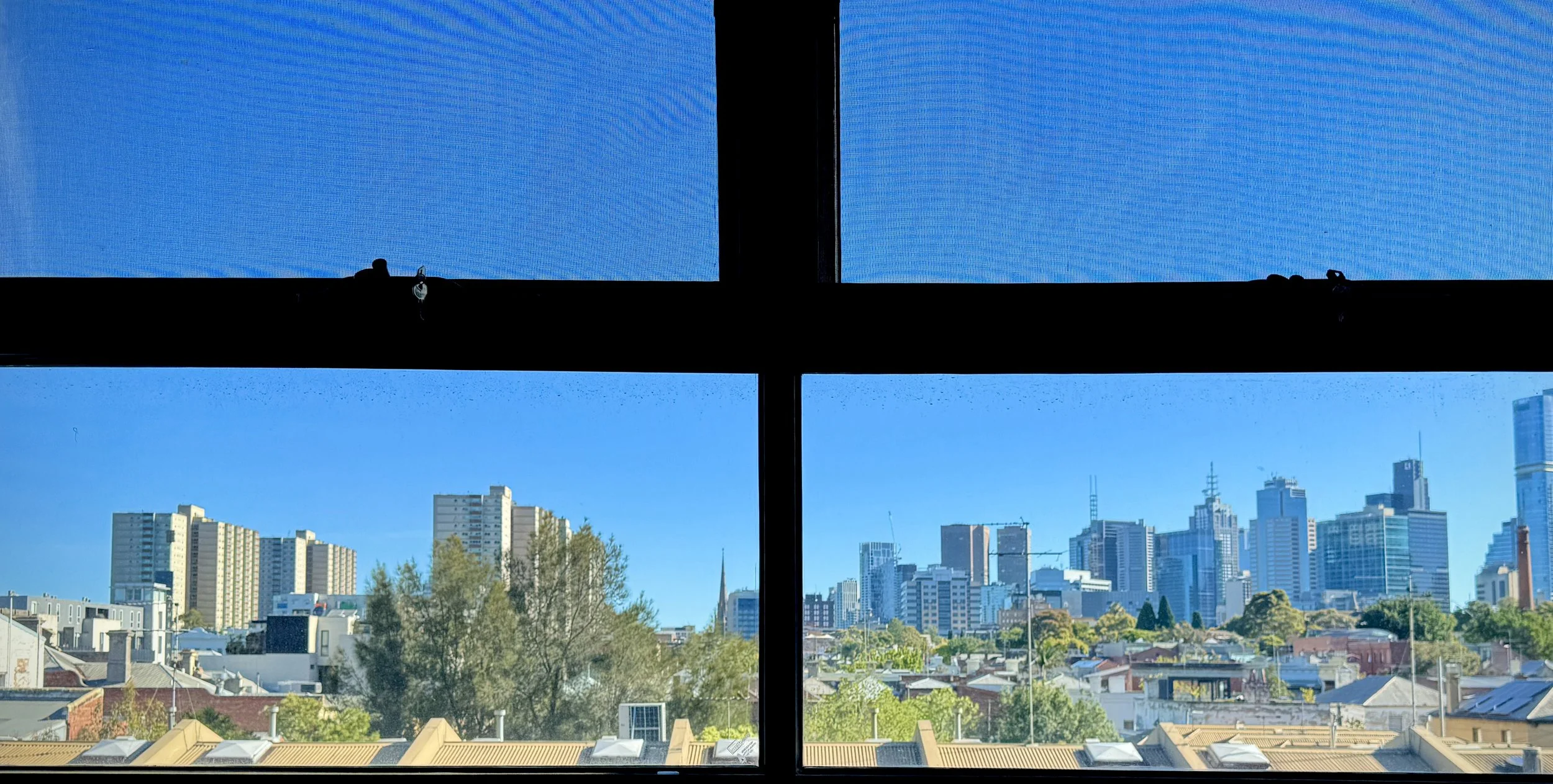 View through a window showing a city skyline with tall buildings, trees, and houses, under a clear blue sky.