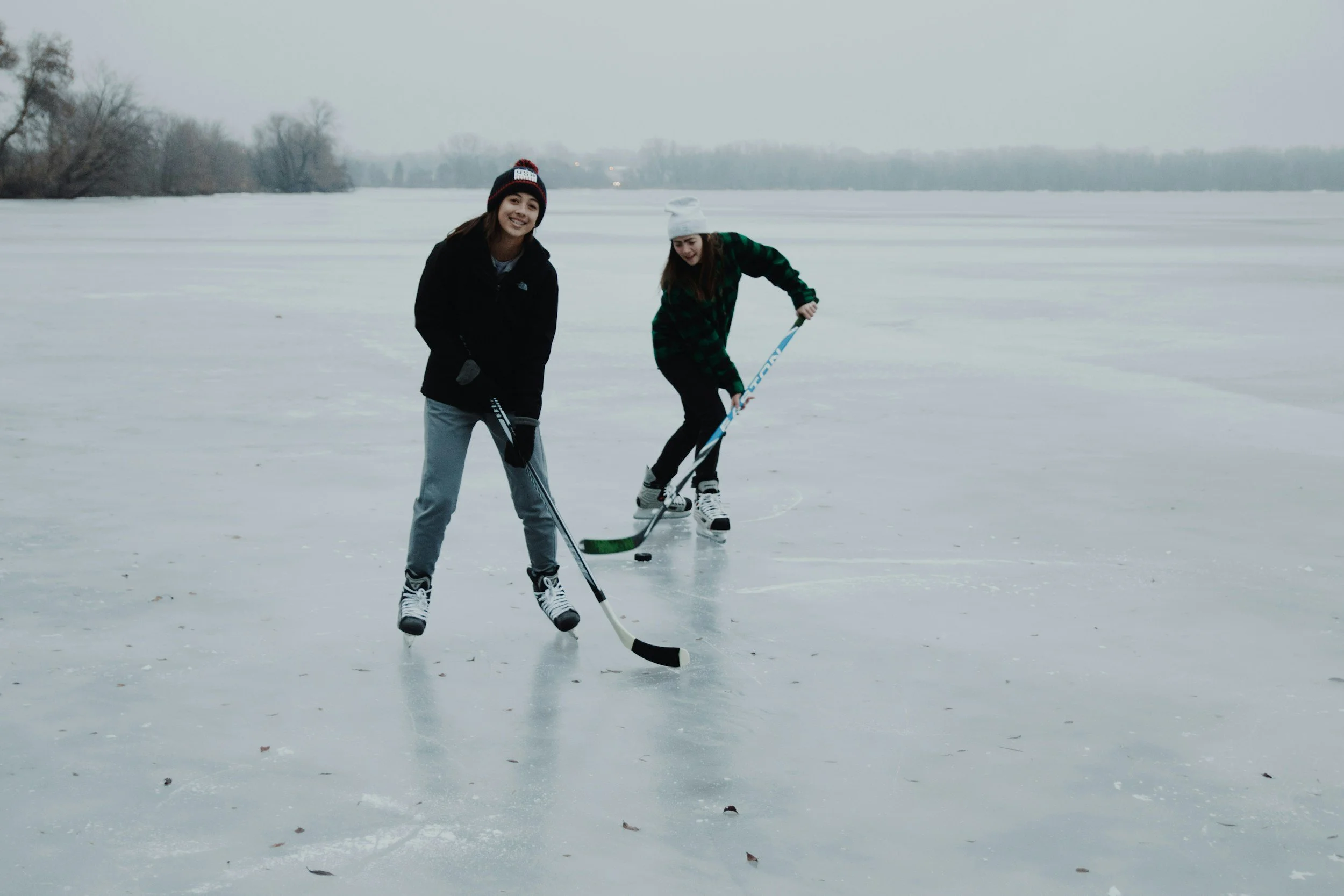 Two young women playing ice hockey on a frozen lake during winter, wearing warm clothing and hats.