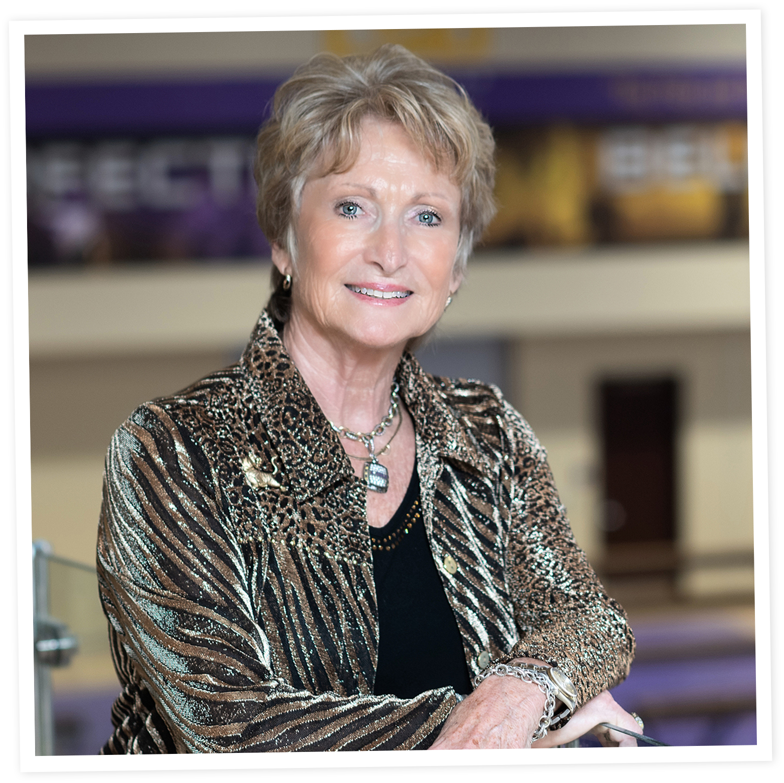 D-D Breaux with short, light brown hair and blue eyes smiling at the camera, wearing a leopard-print jacket, necklaces, and jewelry, in an indoor setting.