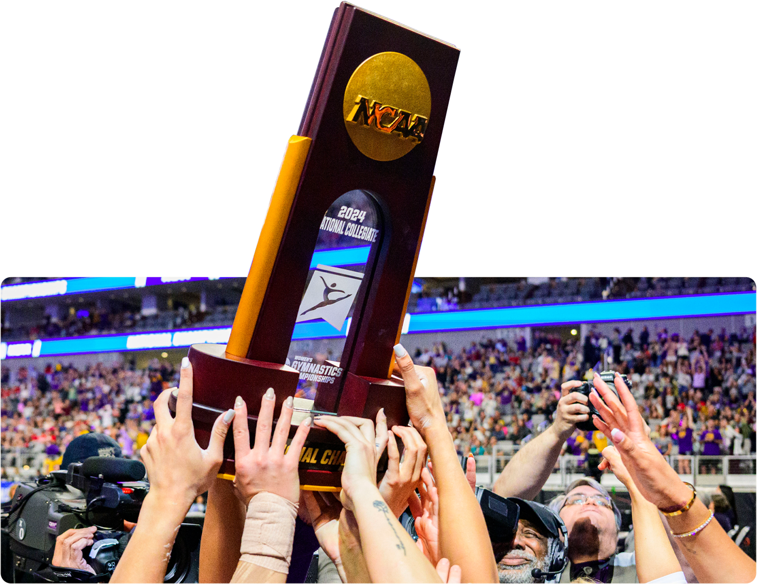 People holding up a large NCAA gymnastics championship trophy at a sports arena, with a crowd in the background.