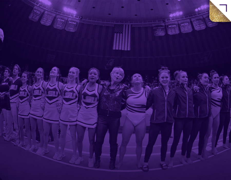 Gymnastics team and coach standing together on a competition floor in an arena with banners hanging from the ceiling.