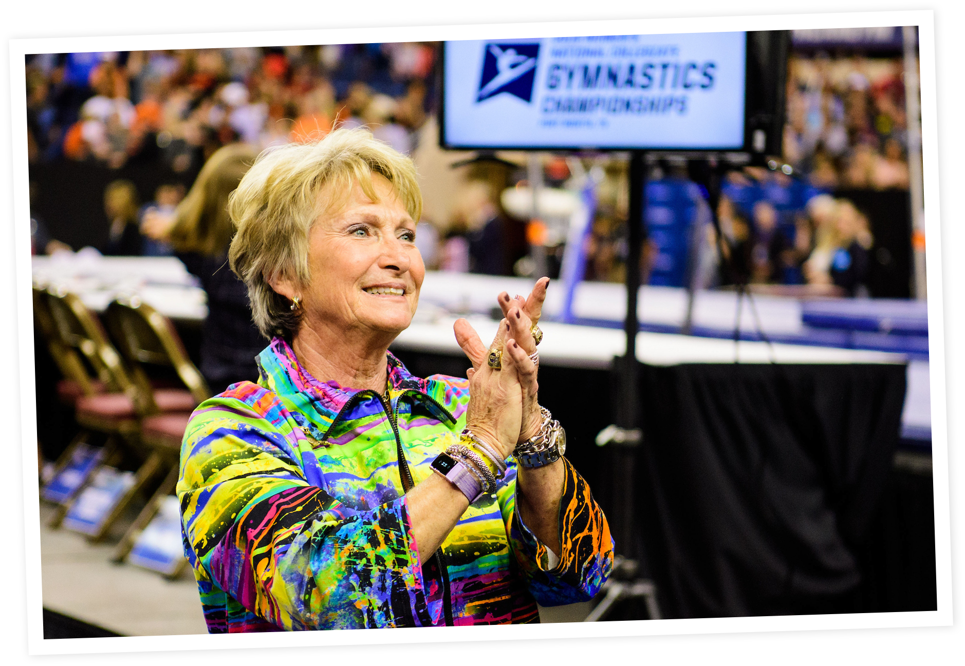 D-D clapping and smiling at a gymnastics competition, with a large screen displaying the competition logo behind her.