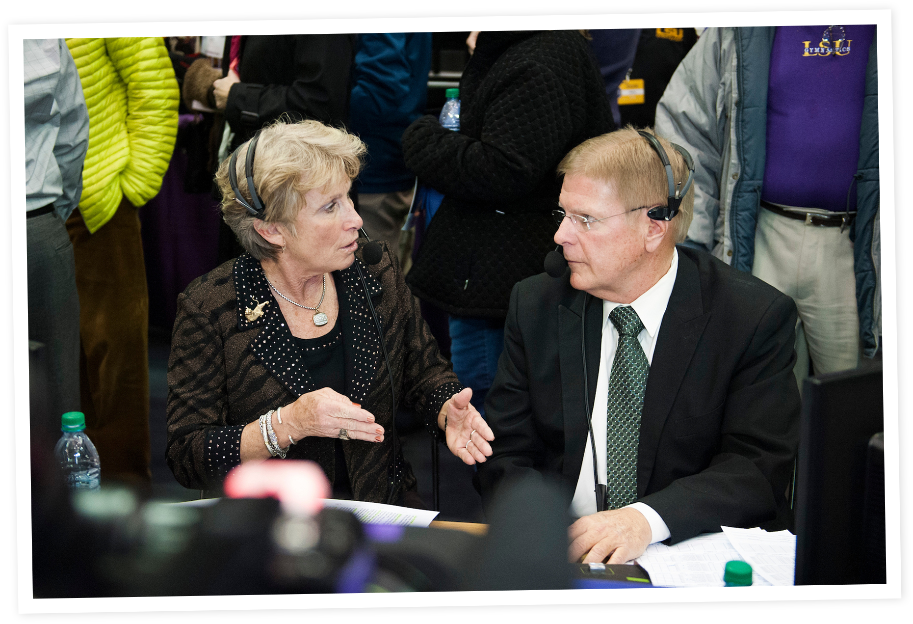 Two people, a woman and a man, are engaged in a conversation with headsets on, seated at a desk with papers and water bottles. The woman is talking animatedly, and the man is listening attentively.