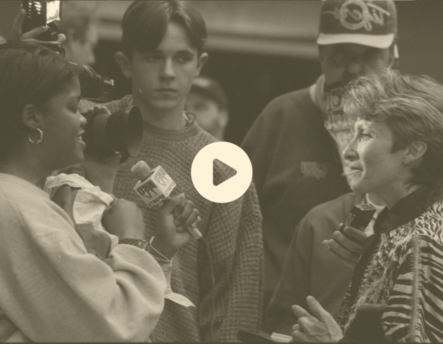 A woman giving an interview with a microphone labeled 'MTV' to another woman, surrounded by a group of people including a young man and a woman with a scarf and glasses, in black and white.