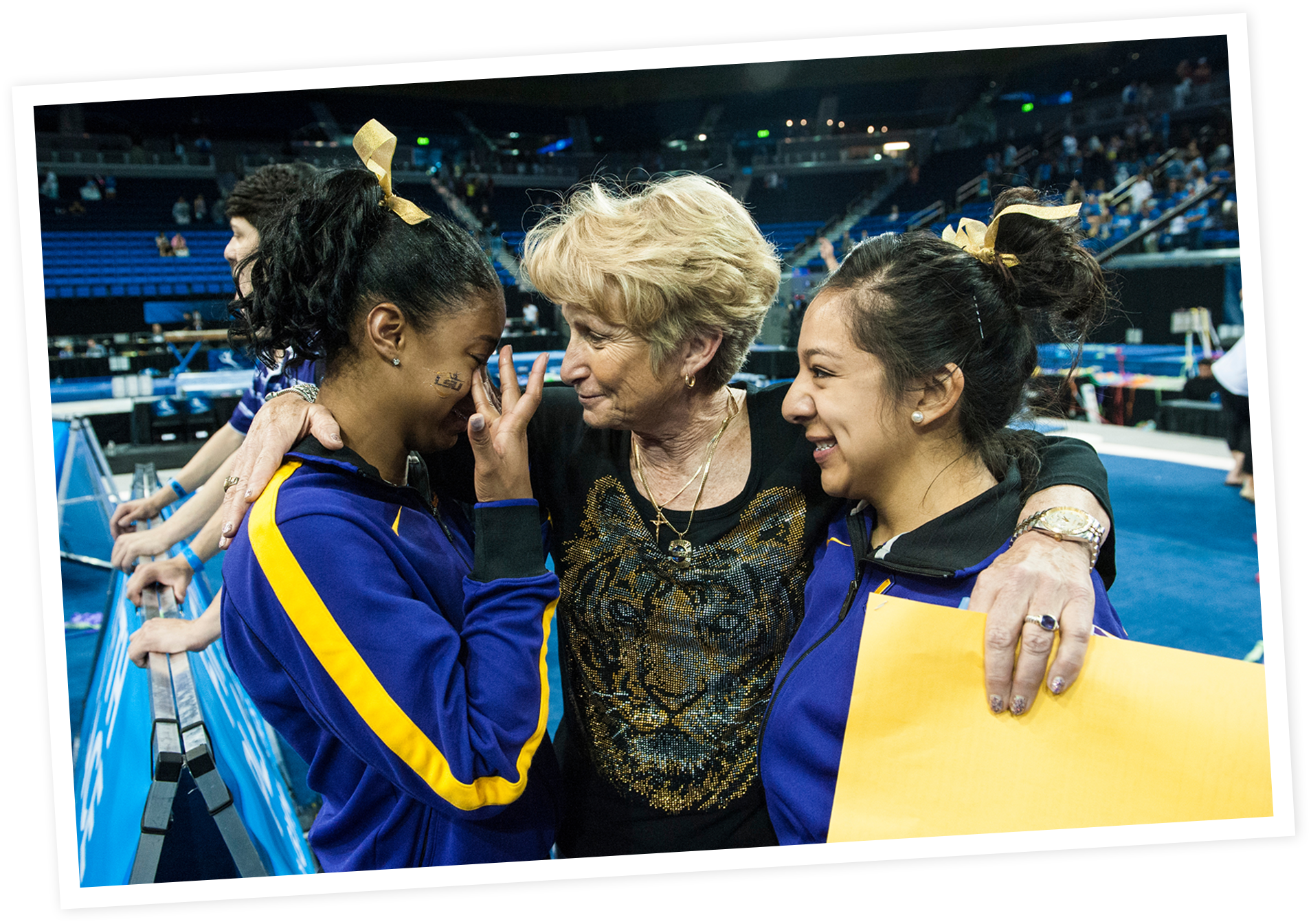 Three women embracing in a sports arena, with two wearing blue and yellow athletic jackets, and the middle woman with short blonde hair and a black shirt with a tiger design, sharing an emotional moment.
