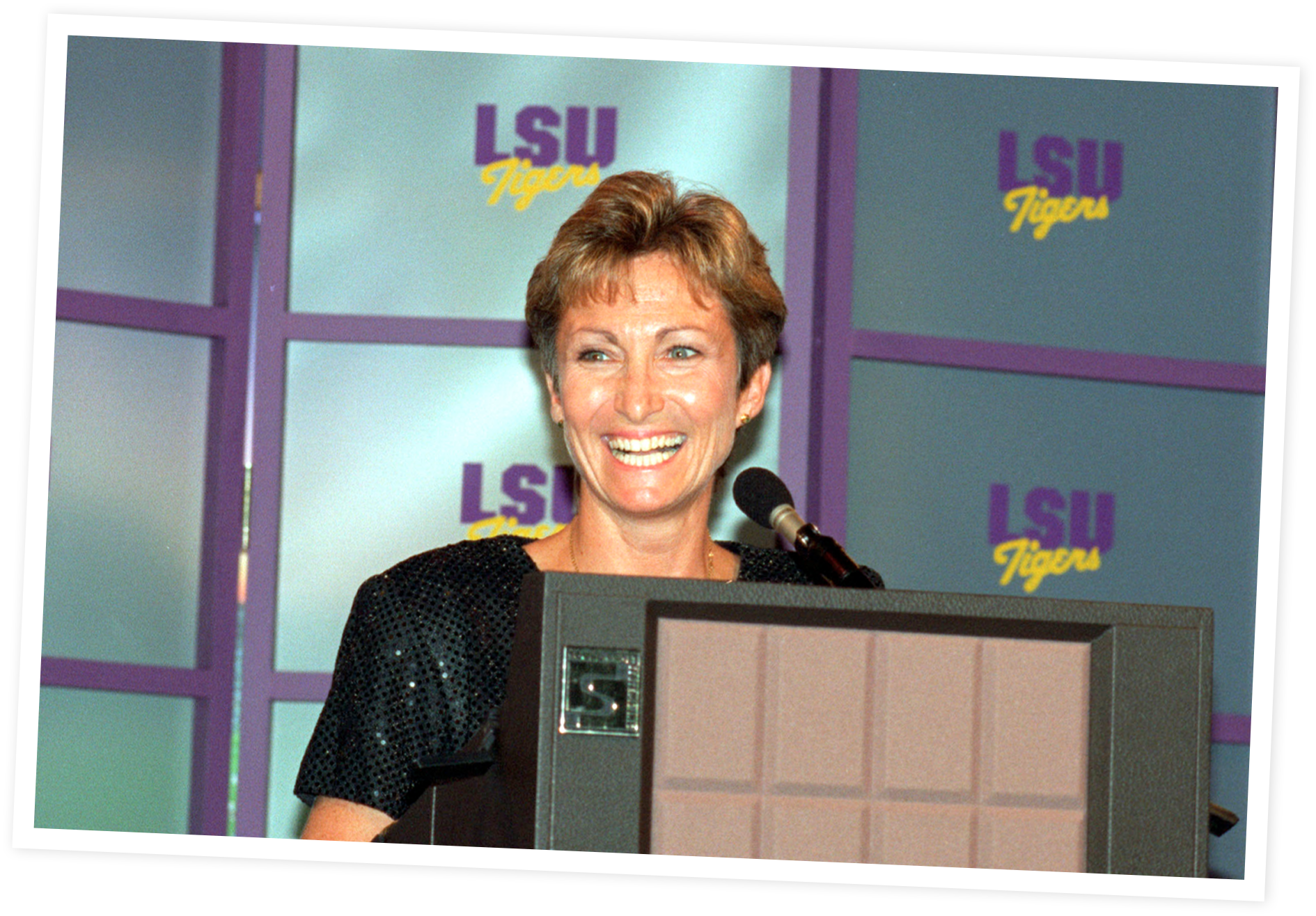 D-D smiling at a podium during a speech or press conference with a backdrop displaying the LSU Tigers logo.
